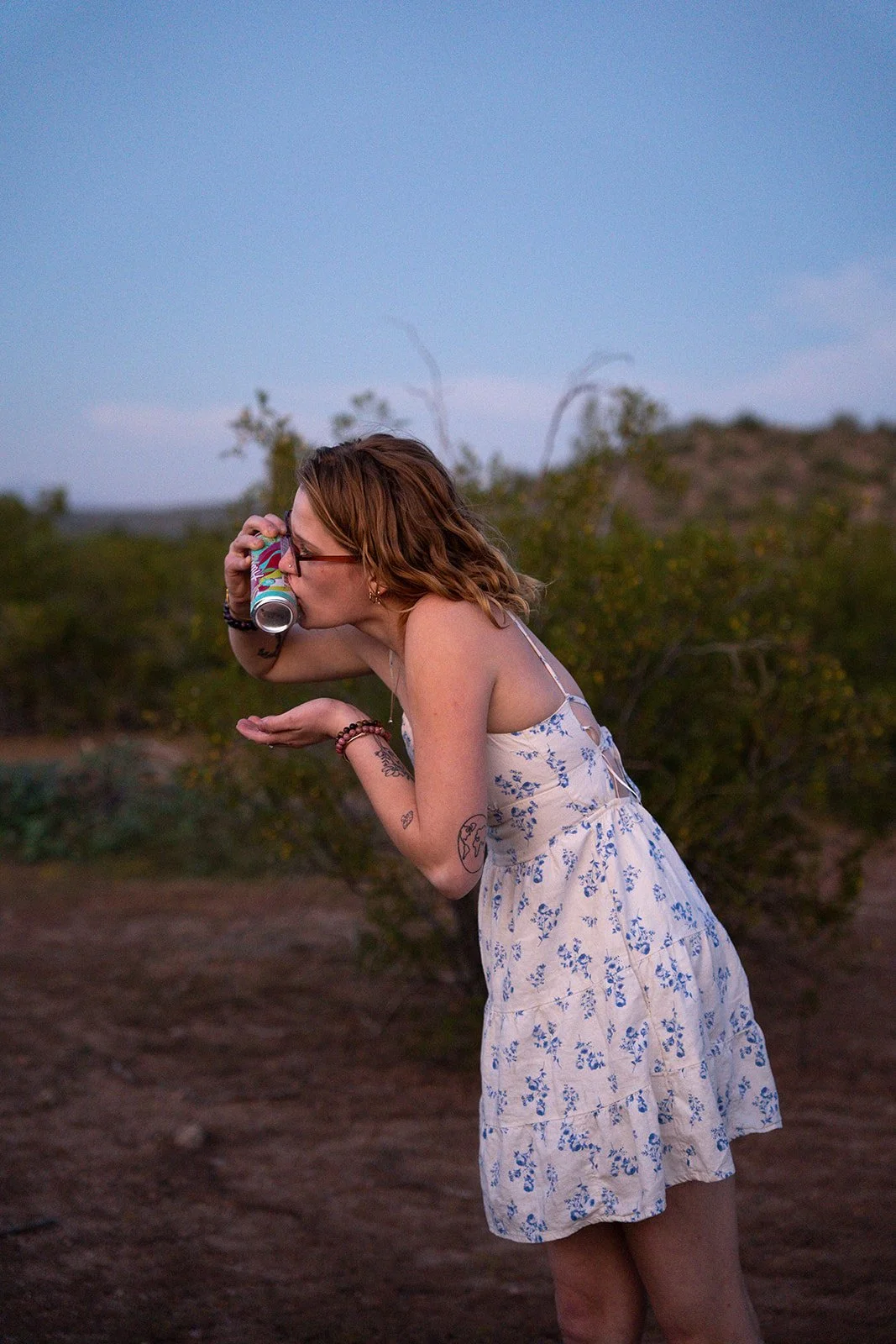 A young woman with tattoos and wearing a white floral dress, standing outdoors during dusk, drinking from a colorful can.