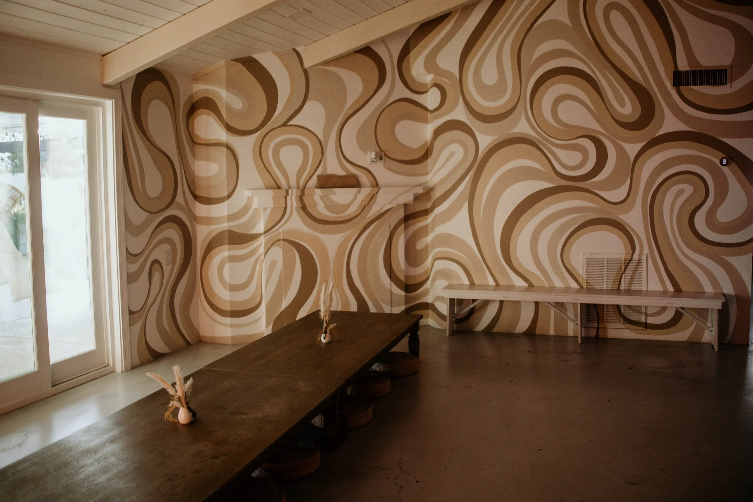 Interior of a room with a bold, abstract, swirling pattern wall in shades of beige, brown, and white. There is a large window with a sliding door on the left, with minimal furniture including a long dark table with small vases on it, a white bench al