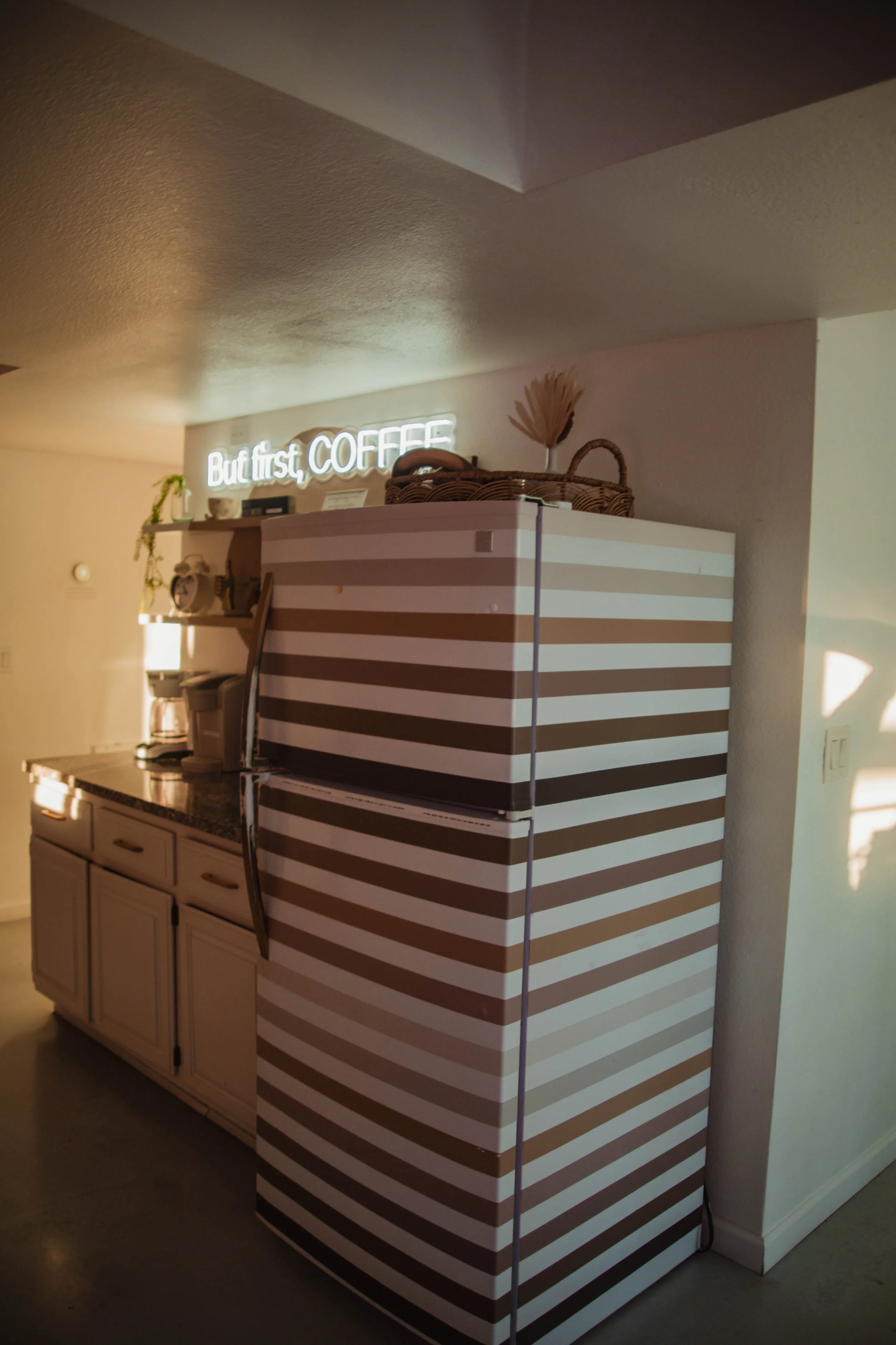 A kitchen corner with a white cabinet, a coffee maker, and a neon sign that says 'But first, COFFEE' on the wall above.