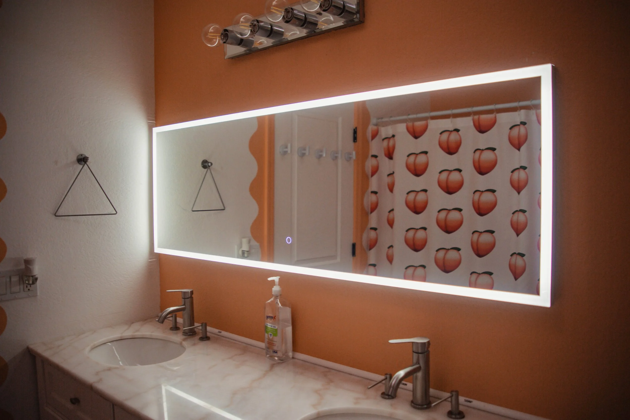 Bathroom mirror with LED lighting above a marble countertop with two sinks, a hand soap dispenser, and a shower curtain with peach and leaf pattern. Decorative hooks on the wall.