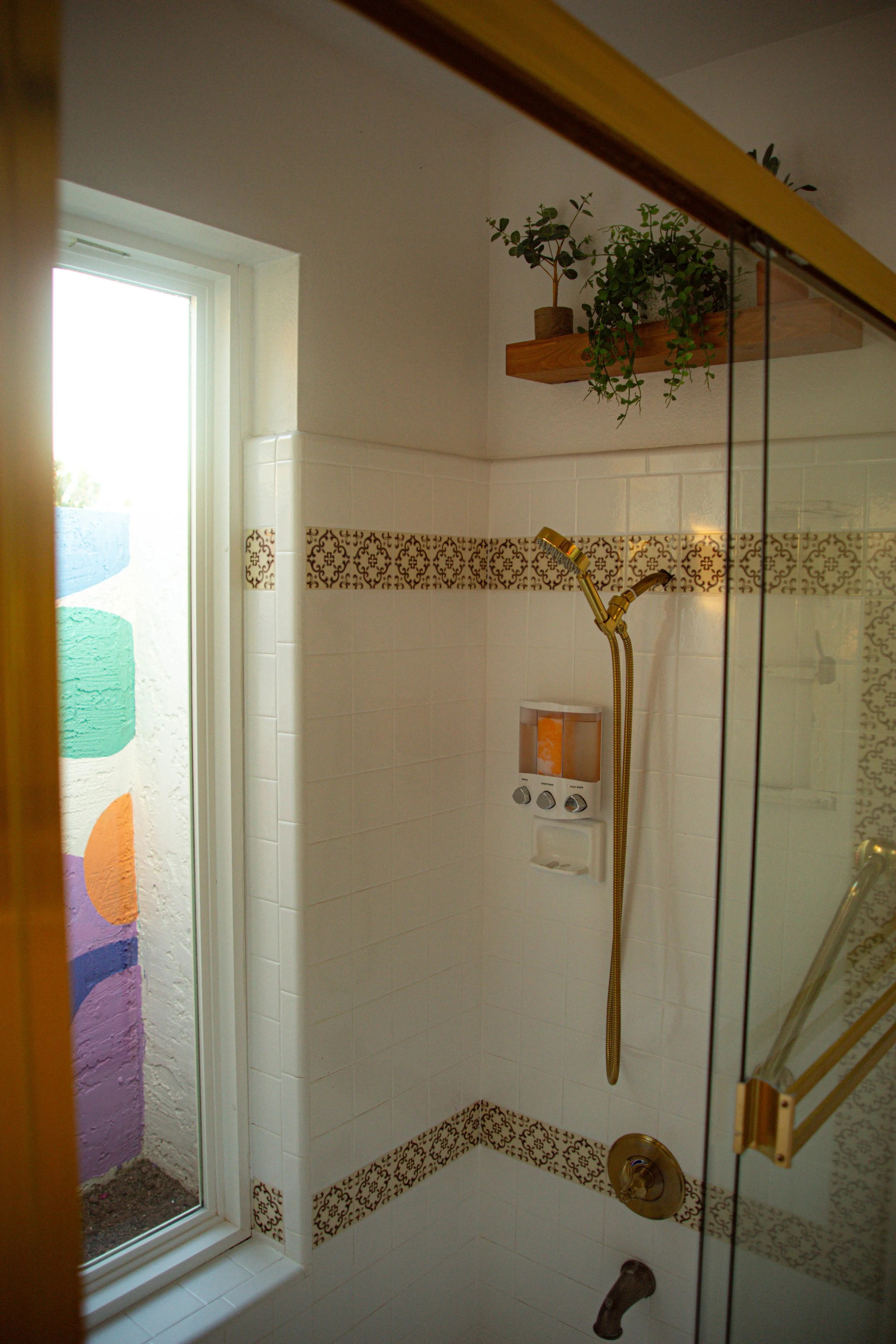 A bathroom shower area with white tile, patterned borders, a gold showerhead, a glass door, and a window with colorful wall art visible outside.