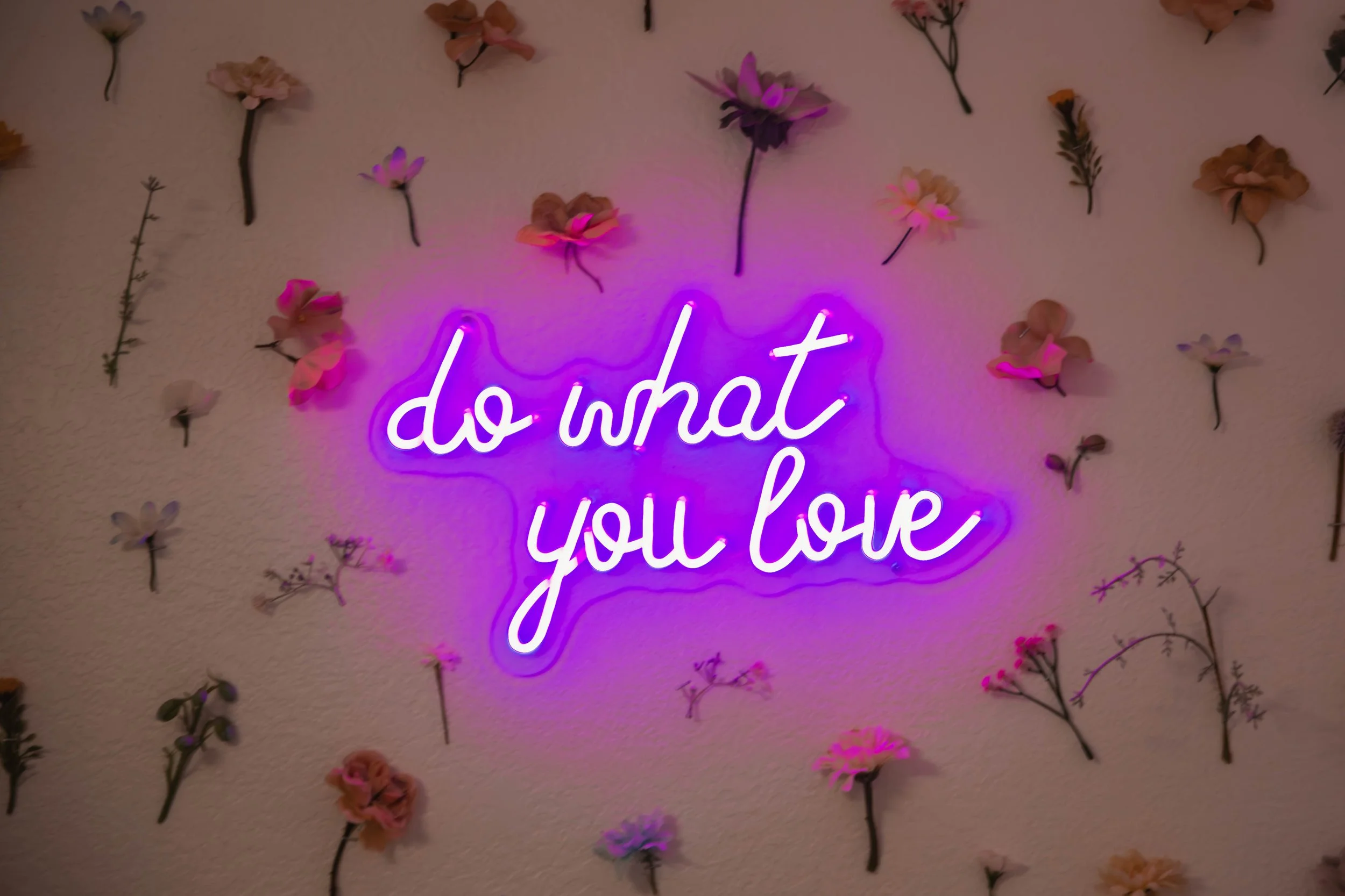 Pink neon sign on a wall that reads 'do what you love' surrounded by various flowers.