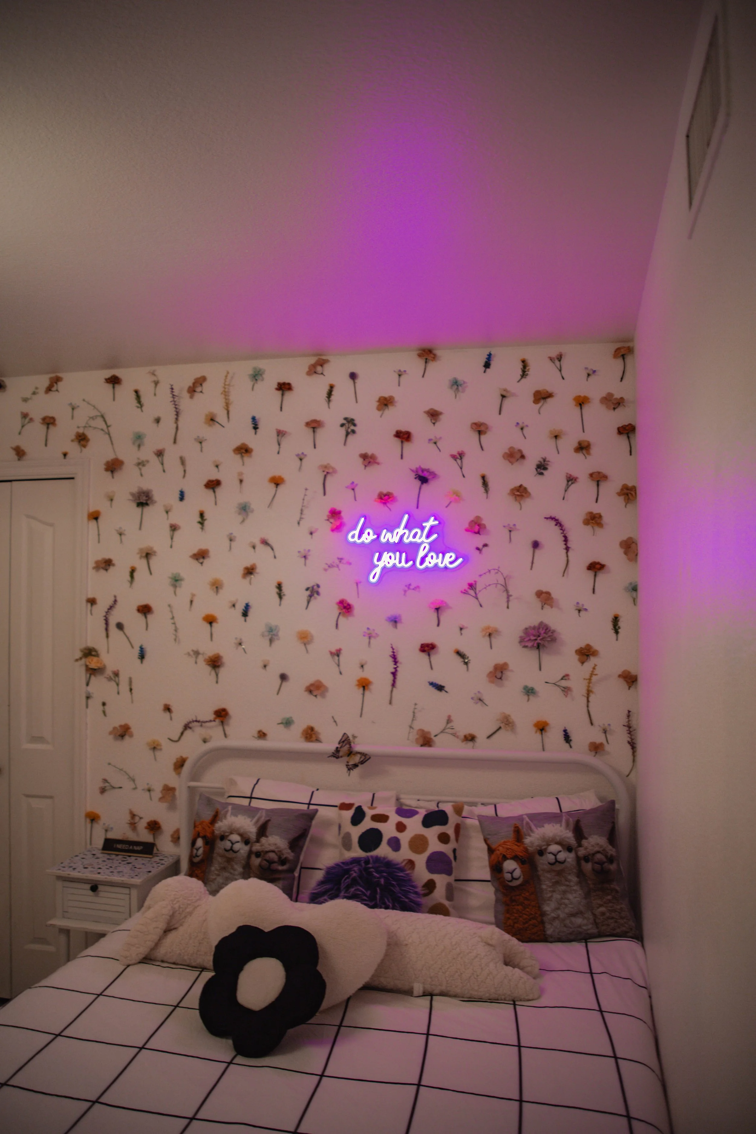 Bedroom with floral wallpaper, neon sign reading 'do what you love,' stuffed animals on bed, and purple and pink lighting