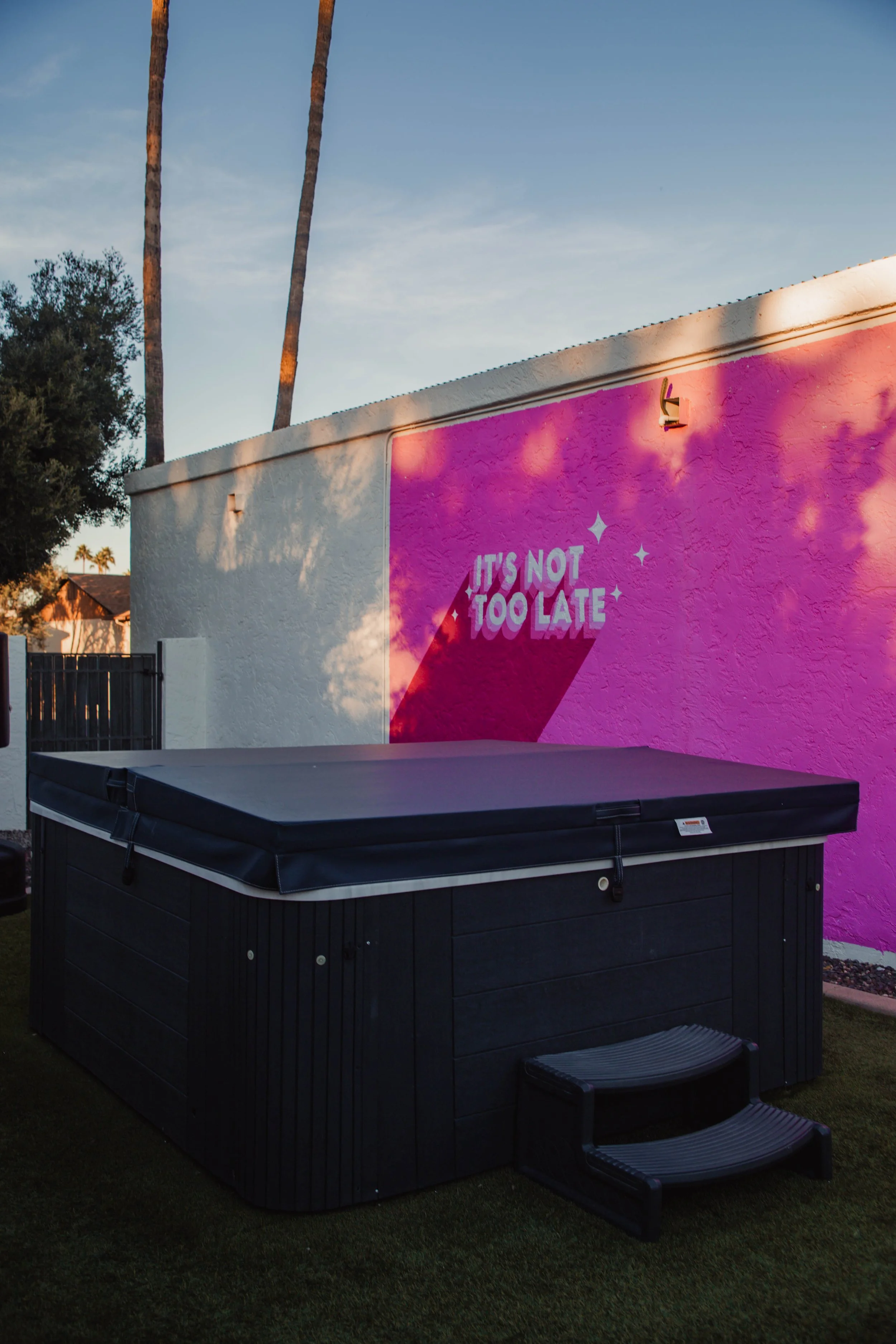A hot tub with a black cover in front of a wall painted pink and white. The wall has text that reads "It's not too late" with a shadow cast on it from nearby trees, and the sky is partly cloudy with palm trees in the background.
