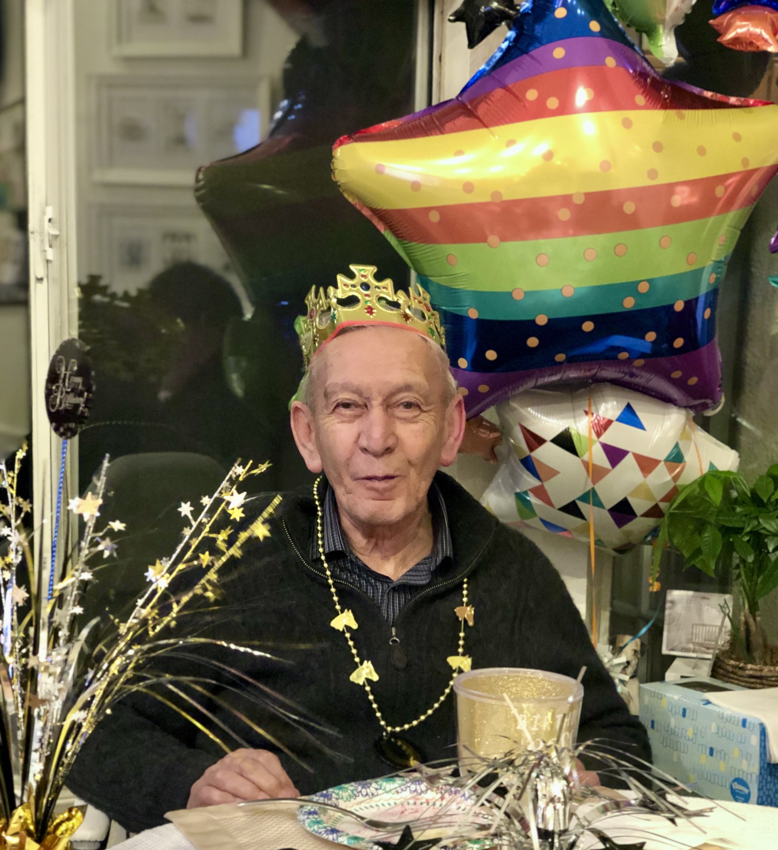 An elderly man wearing a gold paper crown and a gold beaded necklace at a celebration table with colorful balloons, including a large rainbow star-shaped balloon, and festive decorations.