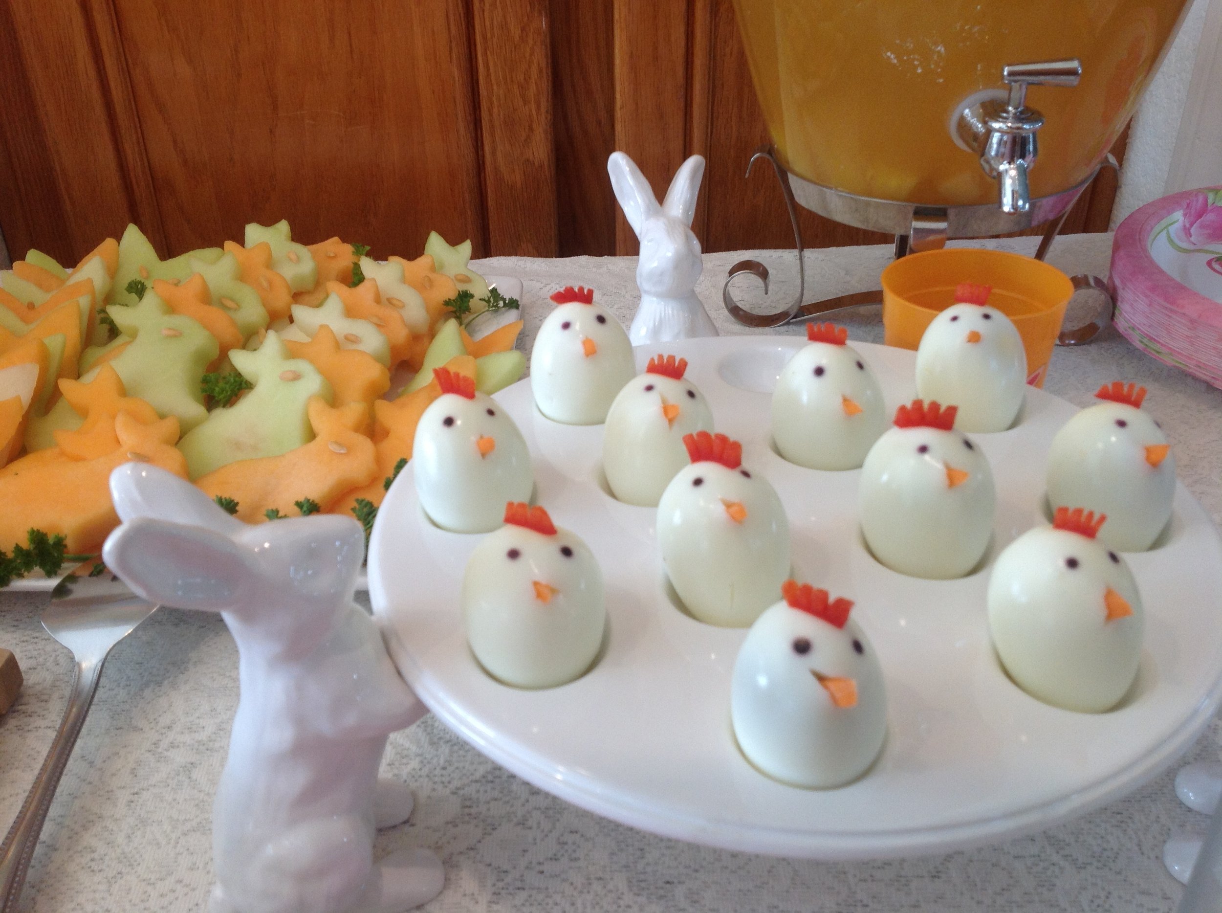 Decorative Easter-themed treats featuring deviled eggs decorated as chickens with small orange beaks, red combs, and tiny black eyes, arranged on a white platter. Additionally, there are cut vegetables on a plate, and ceramic bunny and chick figurine