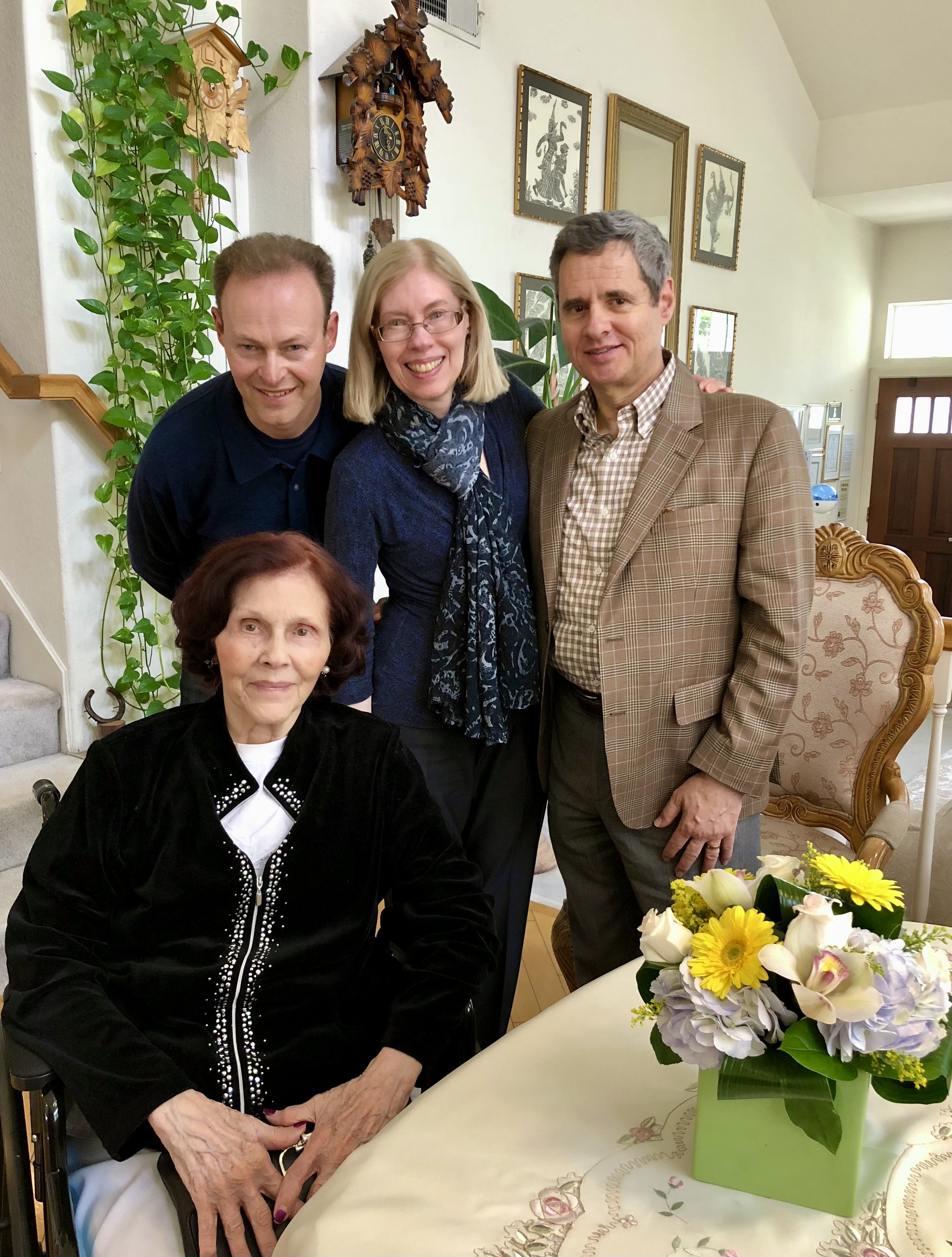 A group of five people gathered in a cozy, well-lit home interior, smiling for the camera. There are three men and two women, with one woman seated in a wheelchair at a dining table adorned with a floral centerpiece. The background features wall art,