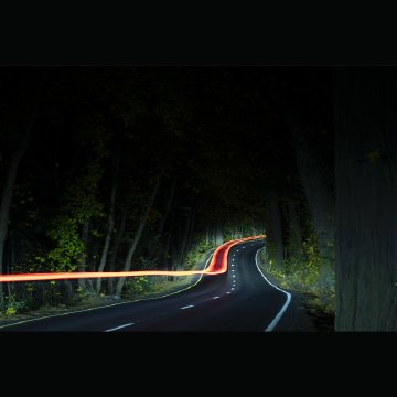 A dark winding road through a forested area at night with light trails from a passing vehicle.