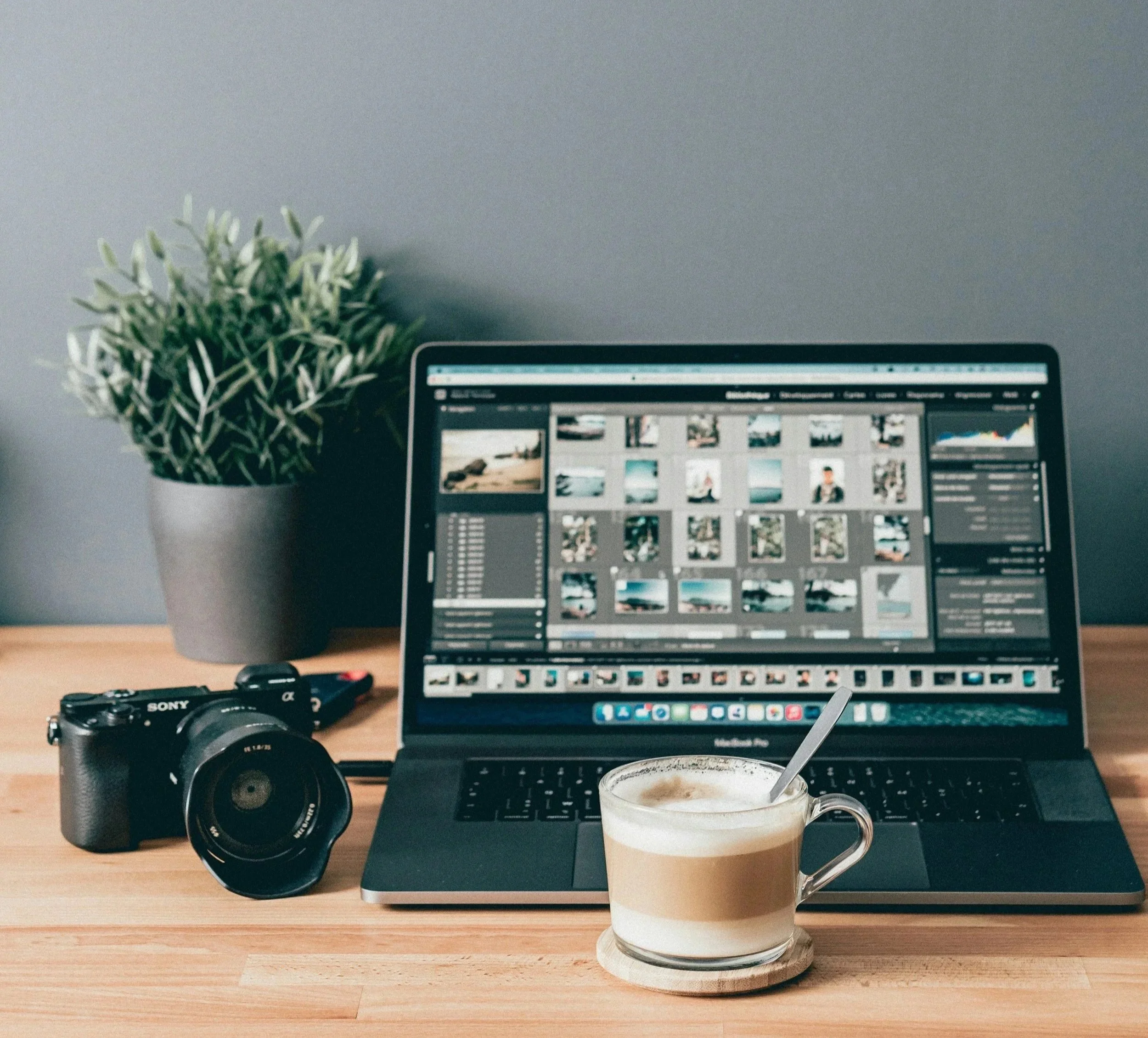A computer on a wooden desk displaying photos, with a camera and a coffee cup in the foreground and a green plant to the left.