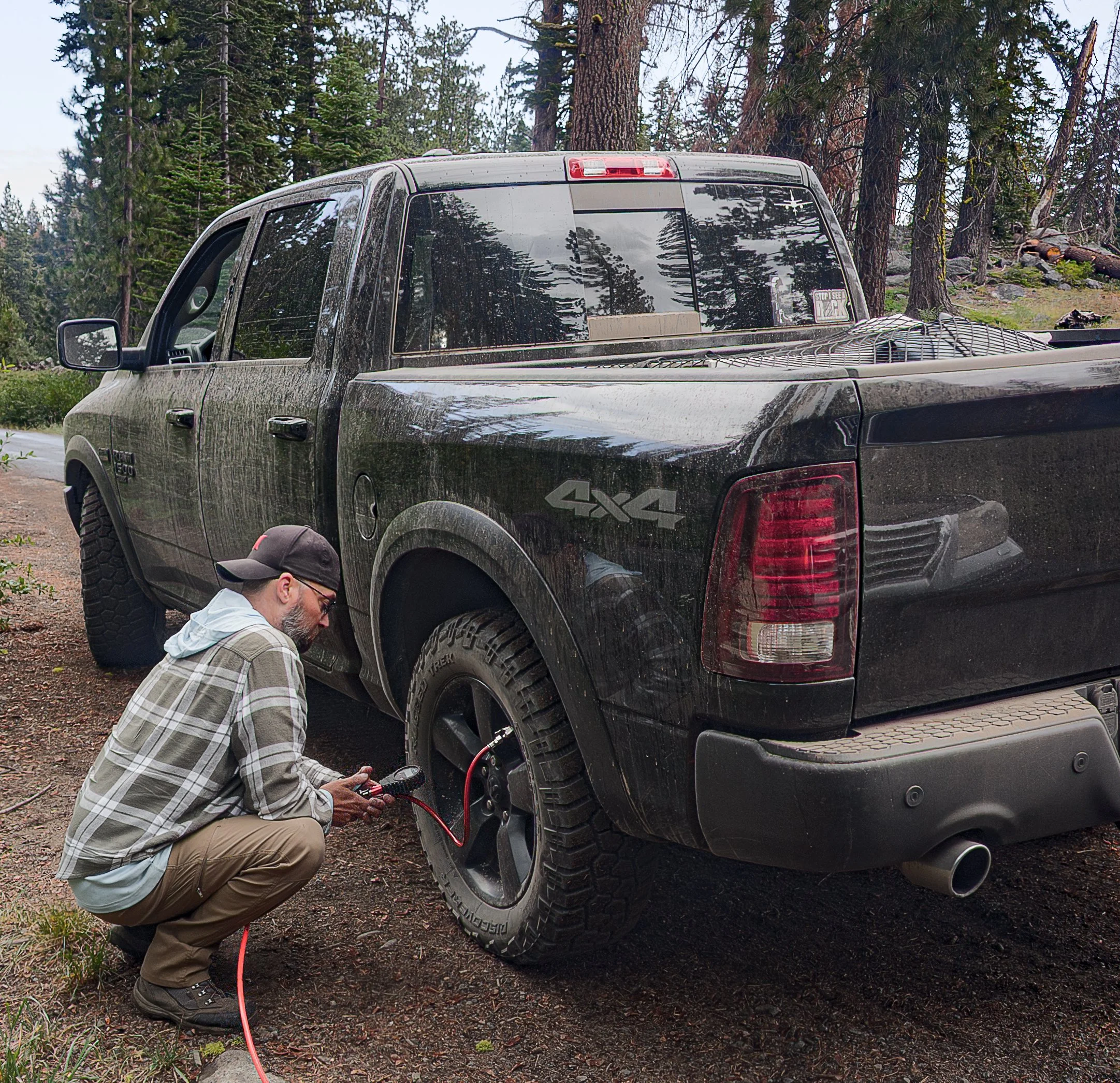 A man kneeling and using an air compressor on a black pickup truck's rear wheel to air up after a private off-road training in a forested area with tall trees.