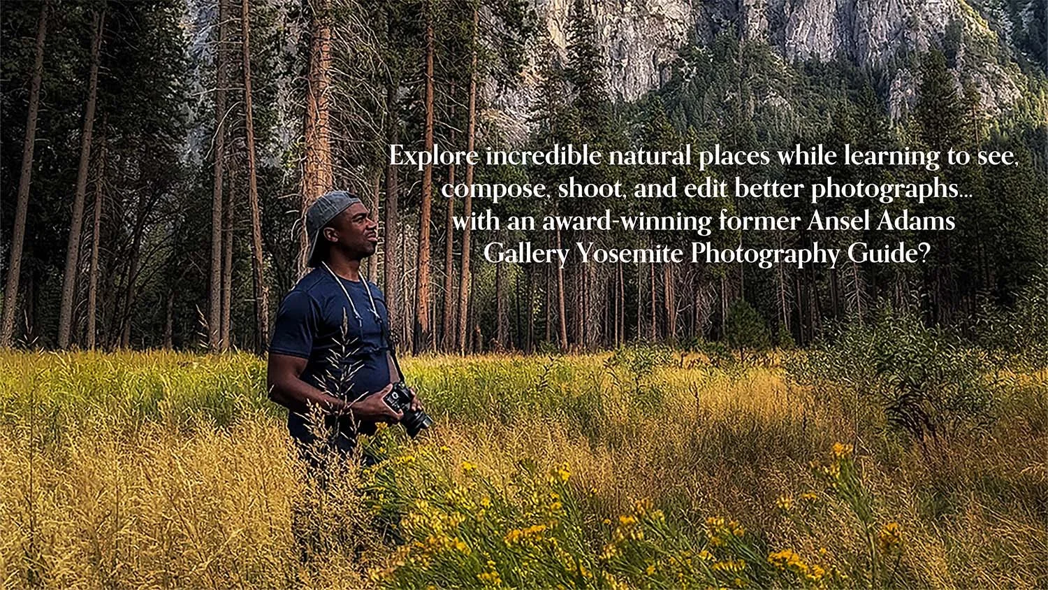 A man on a photography tour with a camera around his neck standing in a field of grass and wildflowers, with a forest of tall trees and rocky cliffs in the background.