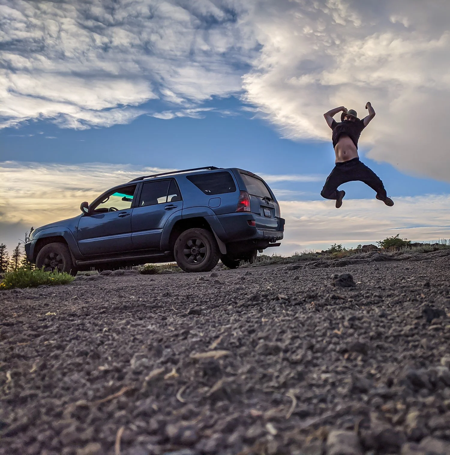 Person jumping beside an off-road vehicle on rocky terrain in Tuolumne County at sunset, with dramatic clouds in the background.