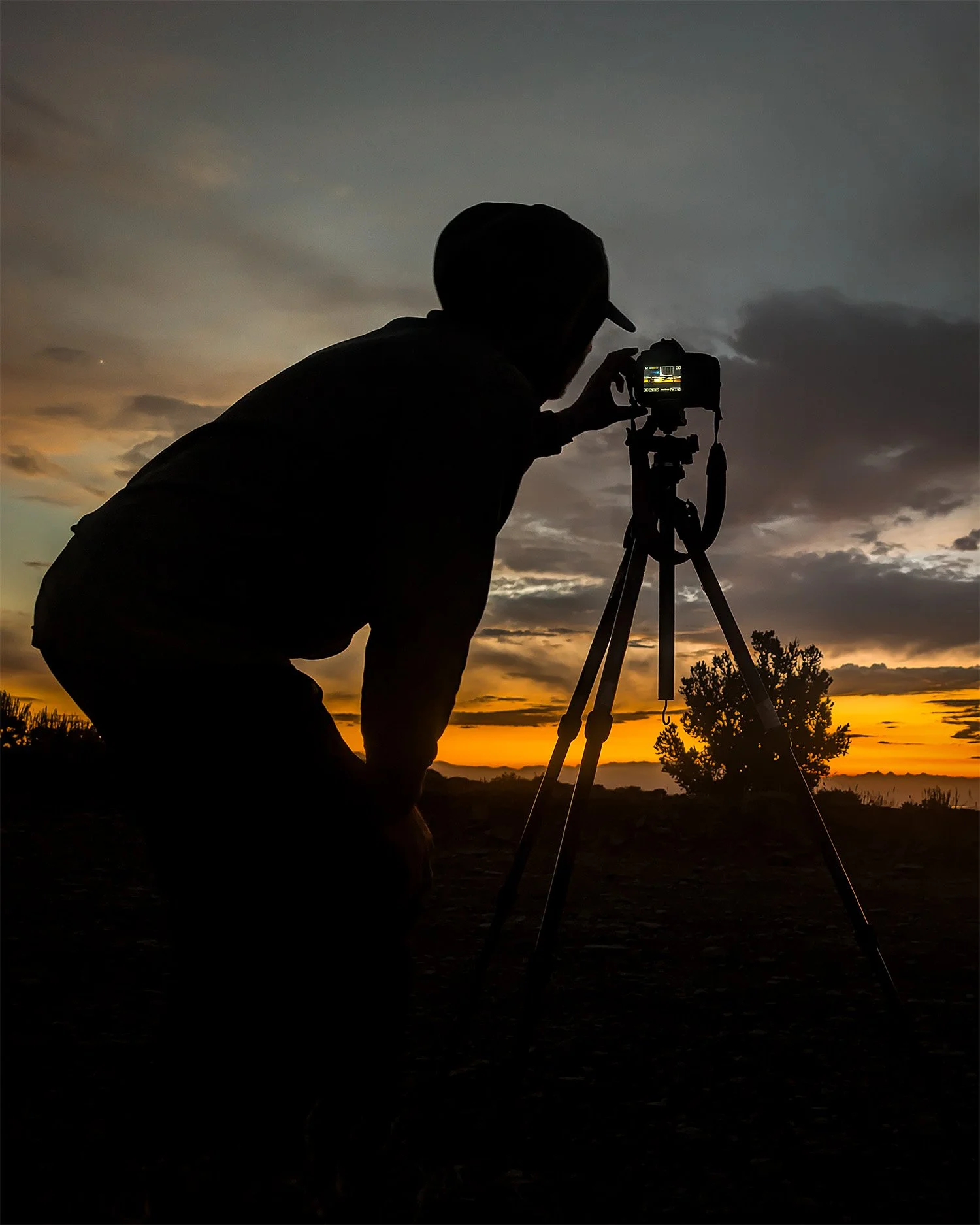 Silhouette of a person wearing a baseball cap, kneeling and adjusting a camera on a tripod during sunset, with a cloudy sky and landscape in the background.