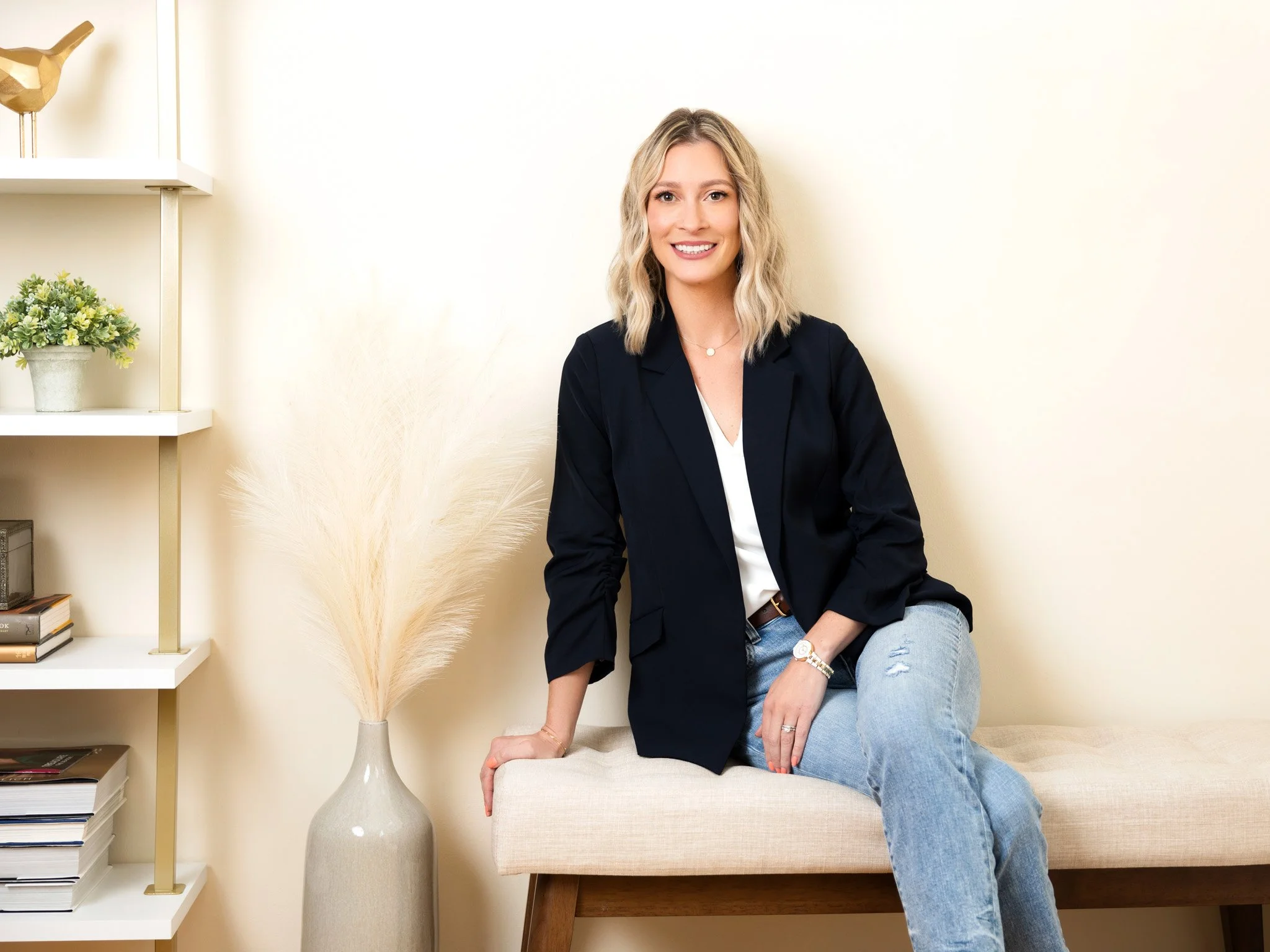 A woman with blonde hair and a friendly smile, wearing a black blazer and ripped jeans, sitting on a beige bench in a bright, modern room with minimalist decor.