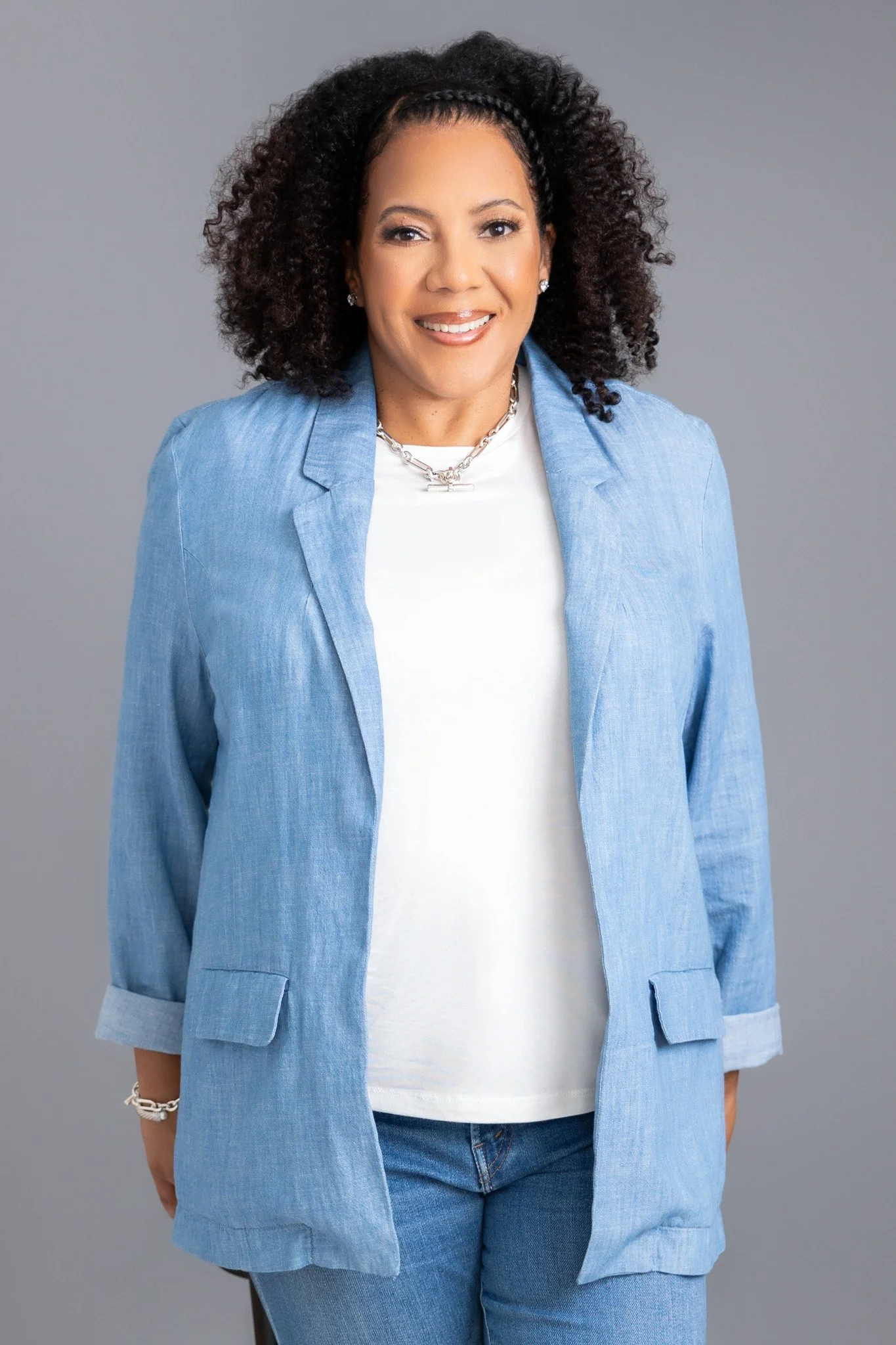 A woman with curly black hair and a hairband, smiling, wearing a light blue blazer over a white shirt, blue jeans, and jewelry, standing against a grey background.