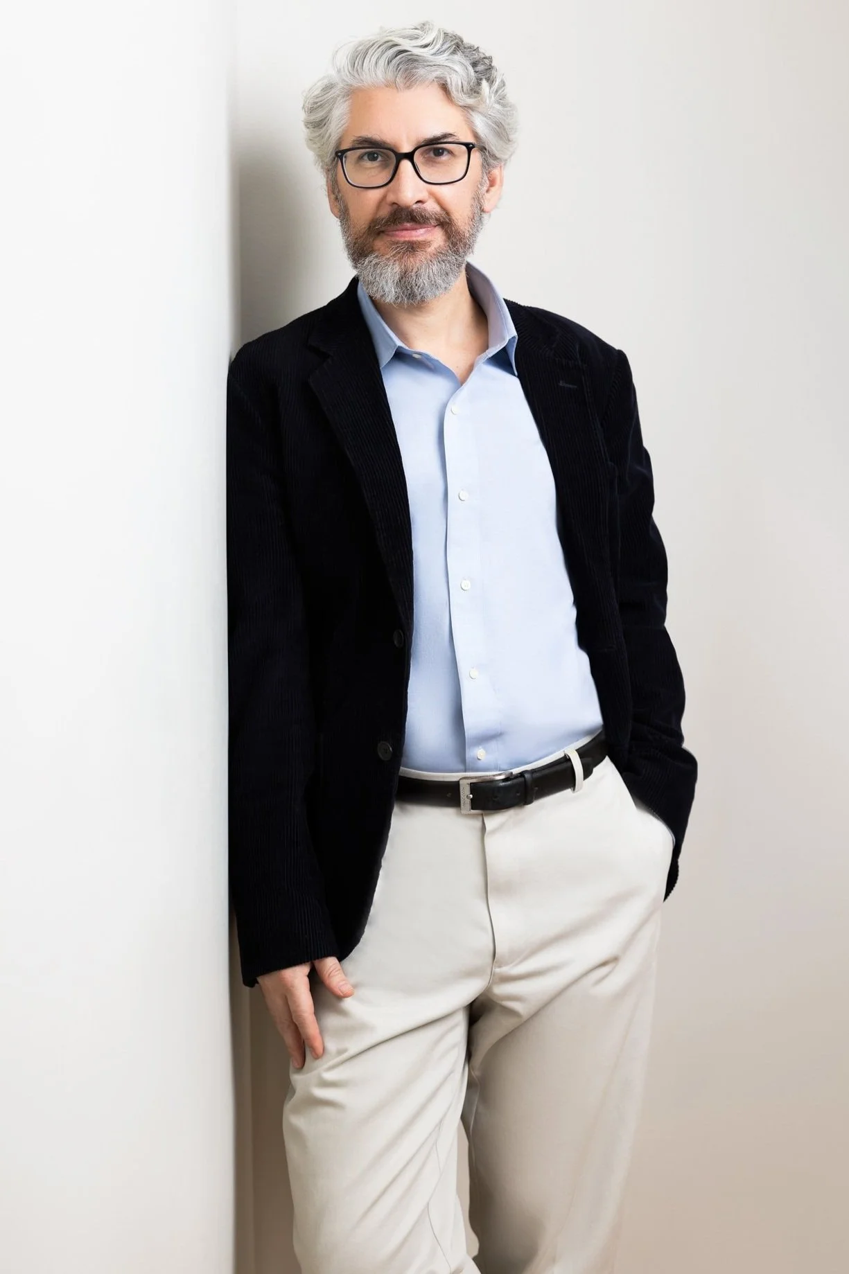 A man with gray hair, beard, and glasses leaning against a white wall, wearing a light blue shirt, dark blazer, and cream-colored pants.