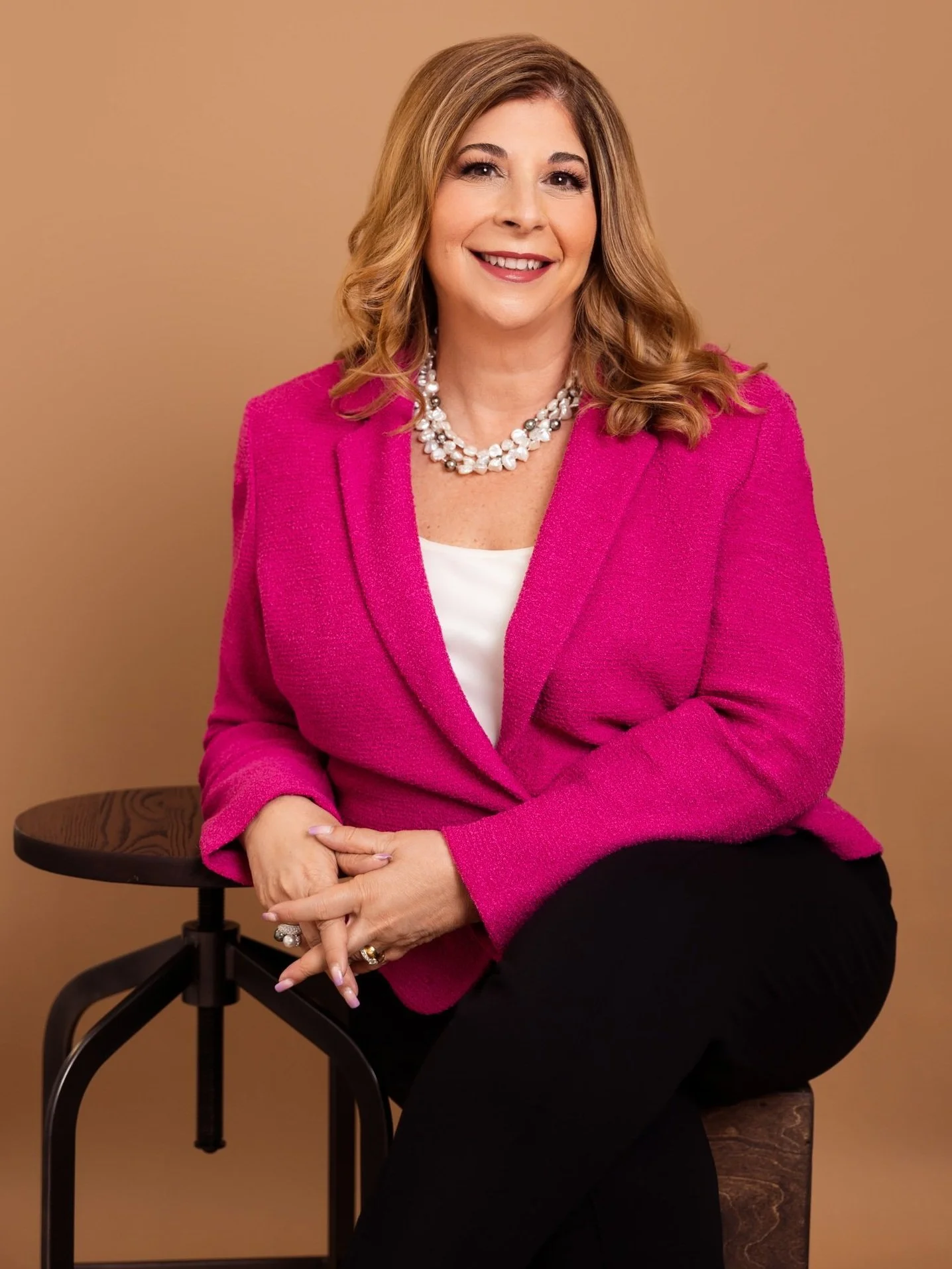 A woman with shoulder-length blonde hair, wearing a bright pink blazer over a white top, black pants, and a pearl necklace, sitting on a stool against a tan background, smiling with her hands clasped.