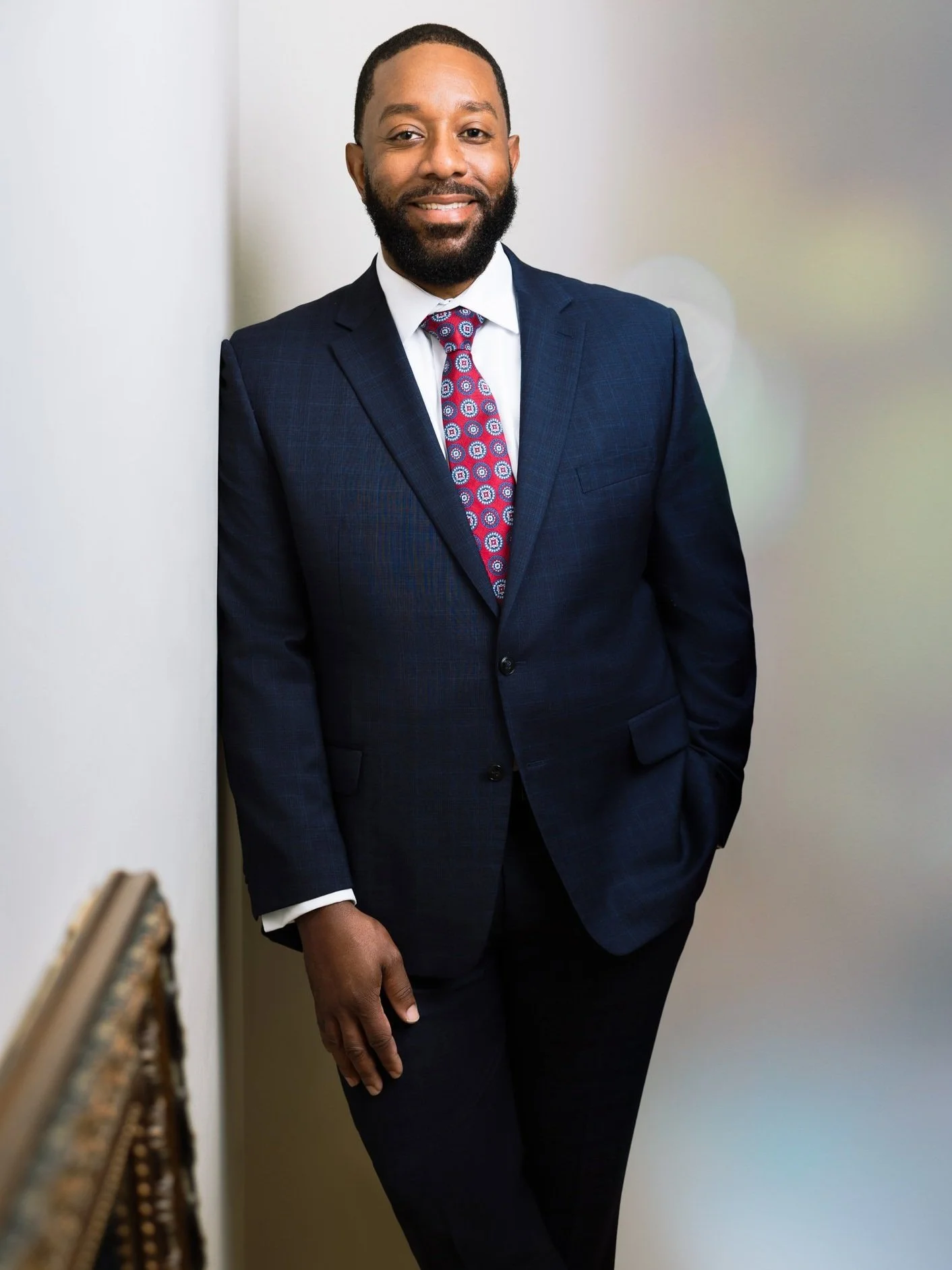 A man in a navy suit with a white shirt and a colorful patterned tie standing against a neutral background, leaning against a wall with his right hand in his pocket and smiling.