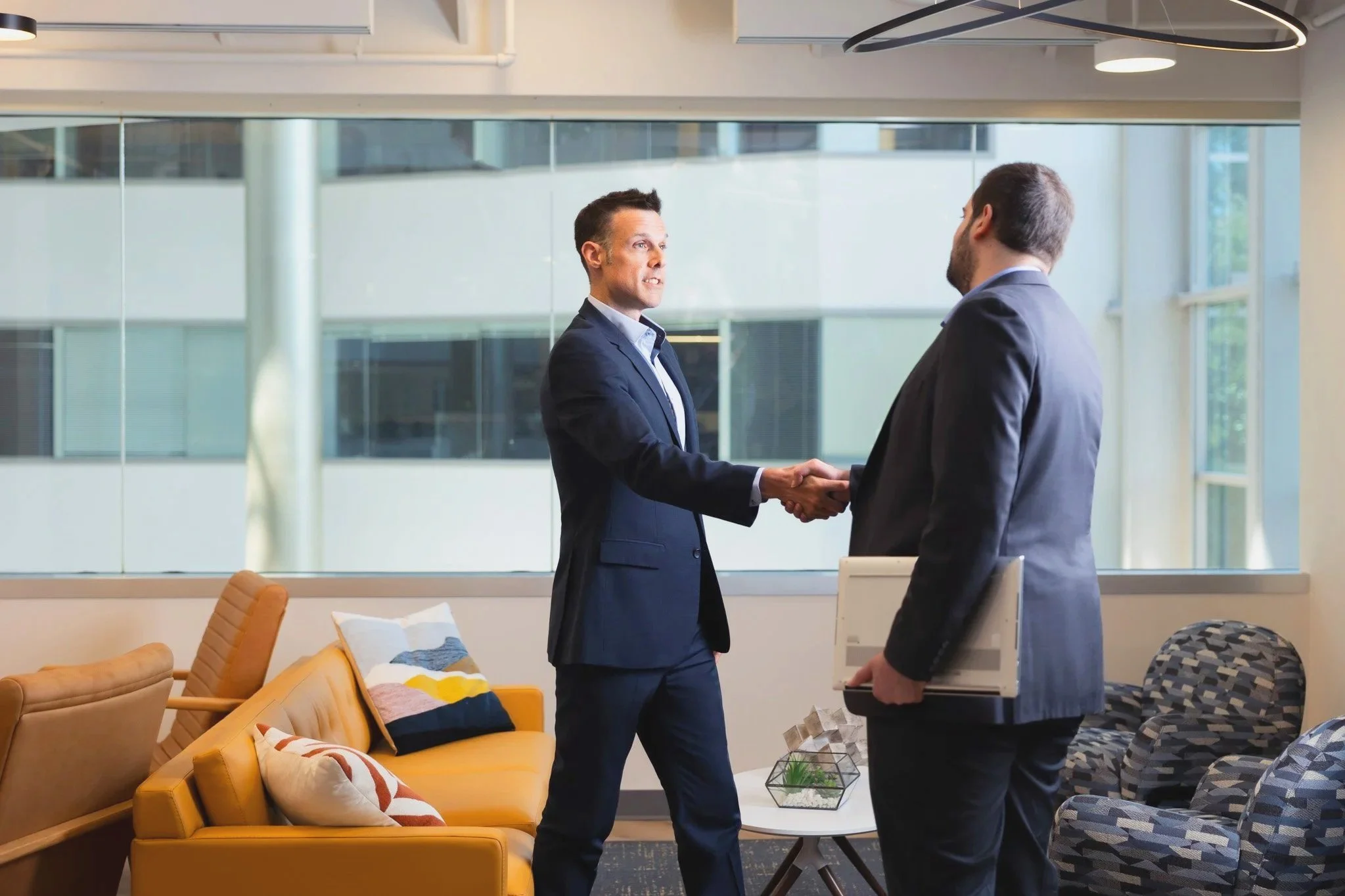 Two men in business suits shake hands in a modern office lounge area with large windows, a yellow sofa, patterned armchairs, and decorative pillows.