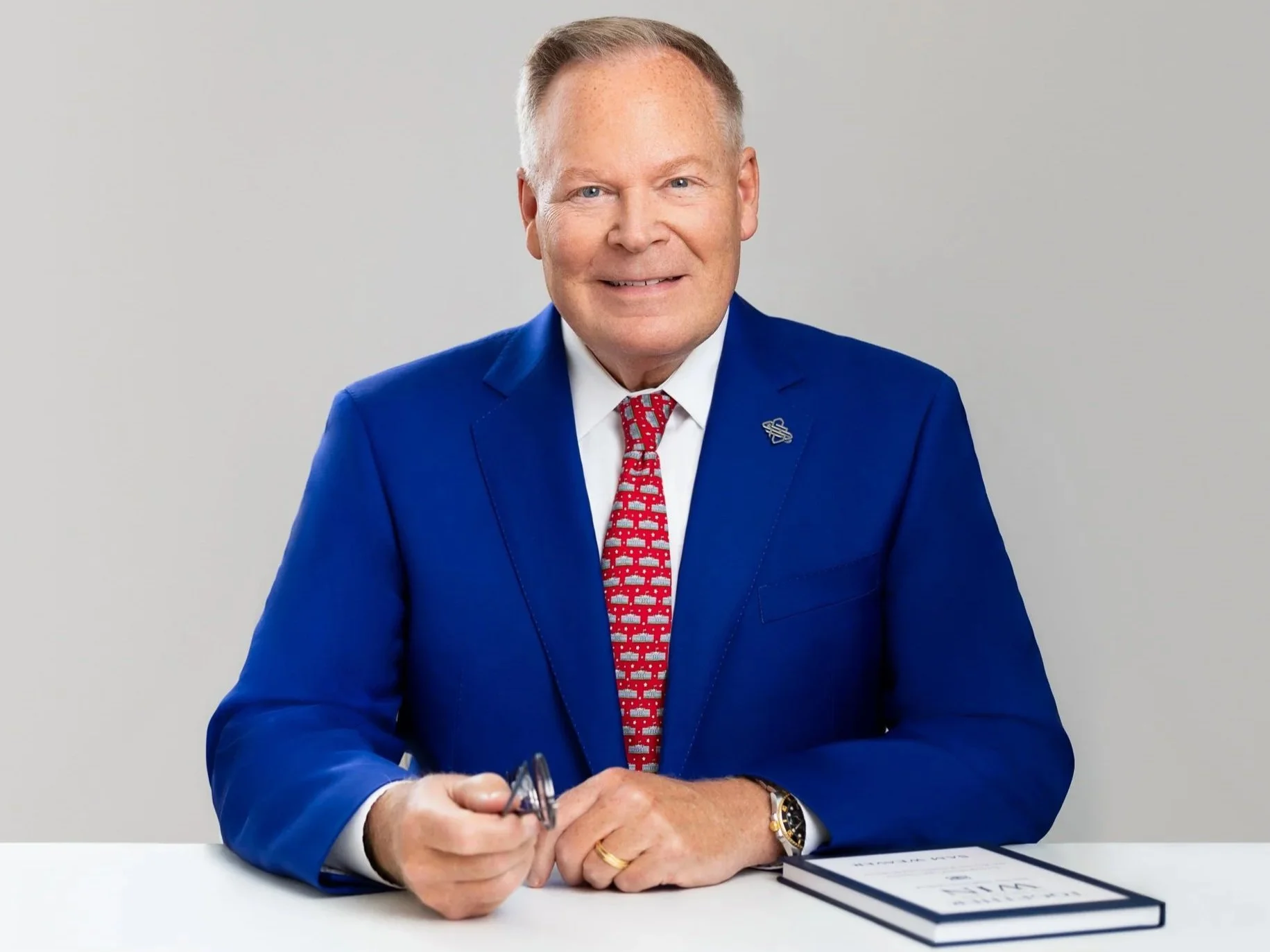 A smiling man holding a pair of glasses, sitting at a white desk with a book in front of him, looking very friendly. He is wearing a bright blue suit and a red tie with a white shirt.