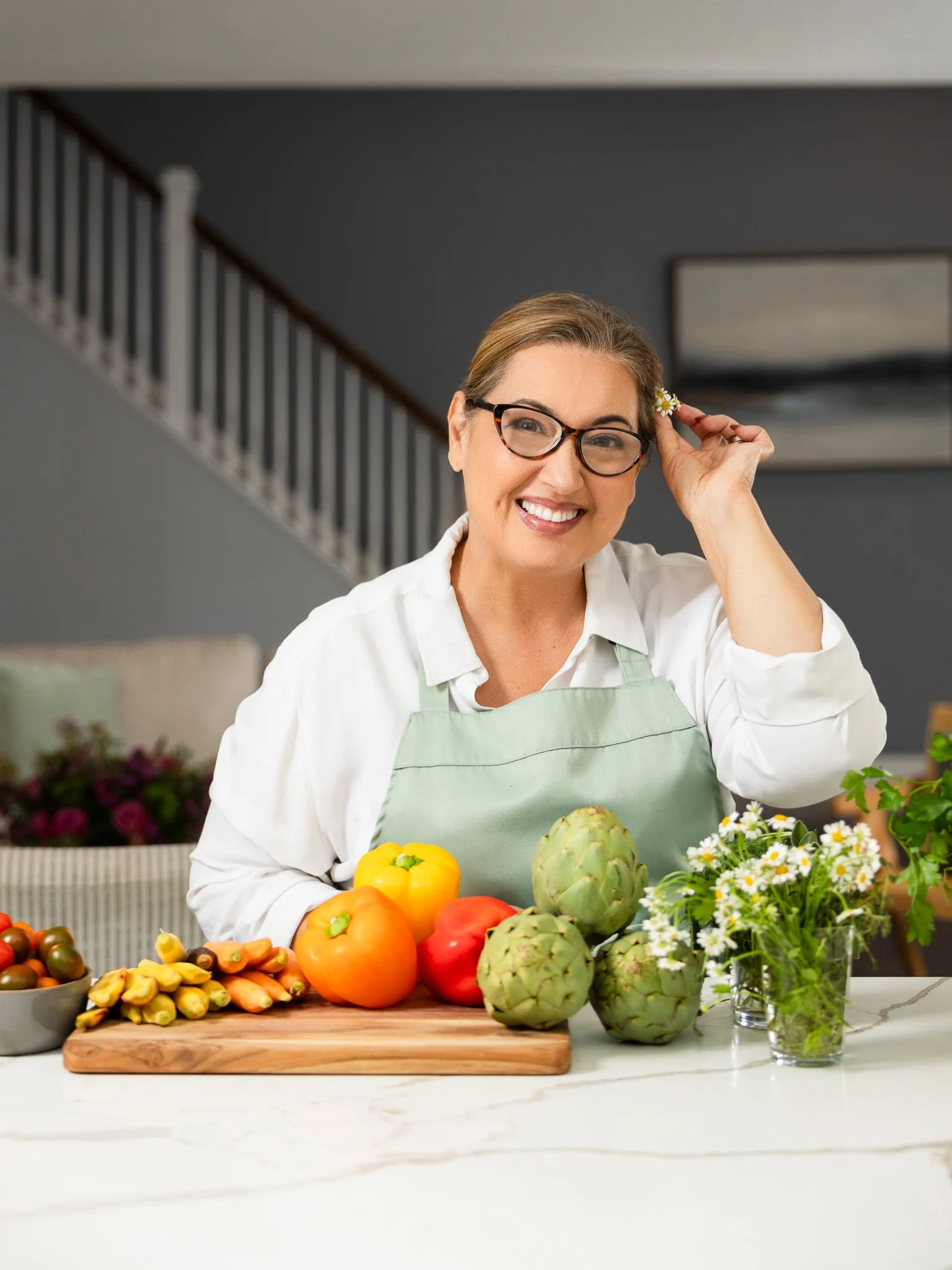 A woman with glasses and a green apron smiling at a kitchen counter with colorful bell peppers, artichokes, carrots, cherry tomatoes, and flowers.