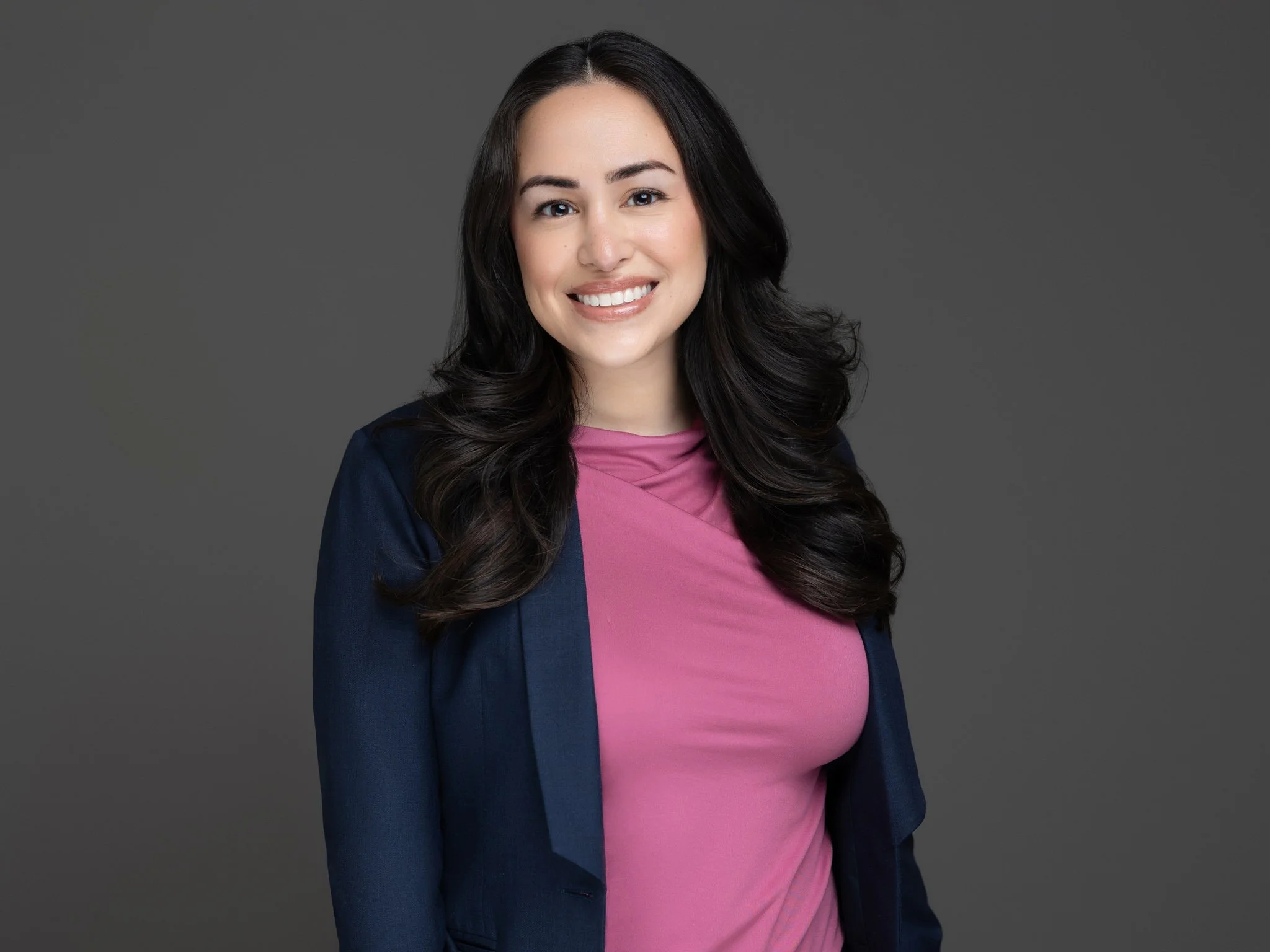 A woman with long dark hair, smiling, wearing a pink top and navy blazer, standing against a plain gray background.