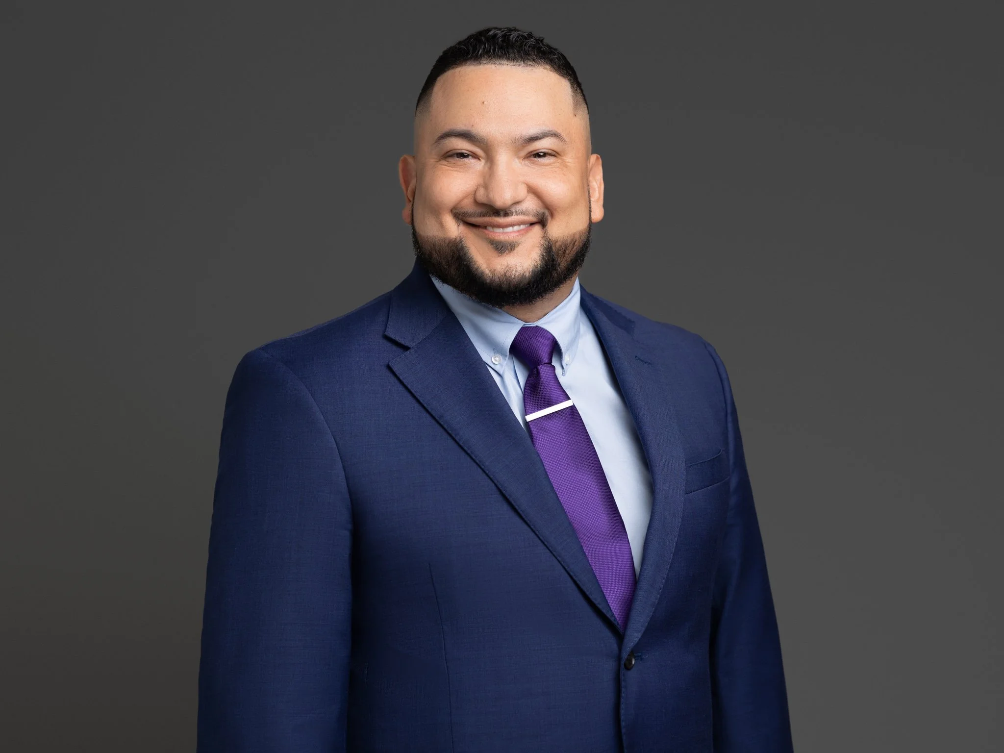 A man with a beard and short dark hair smiling in a blue suit, light blue shirt, purple tie, against a dark gray background.