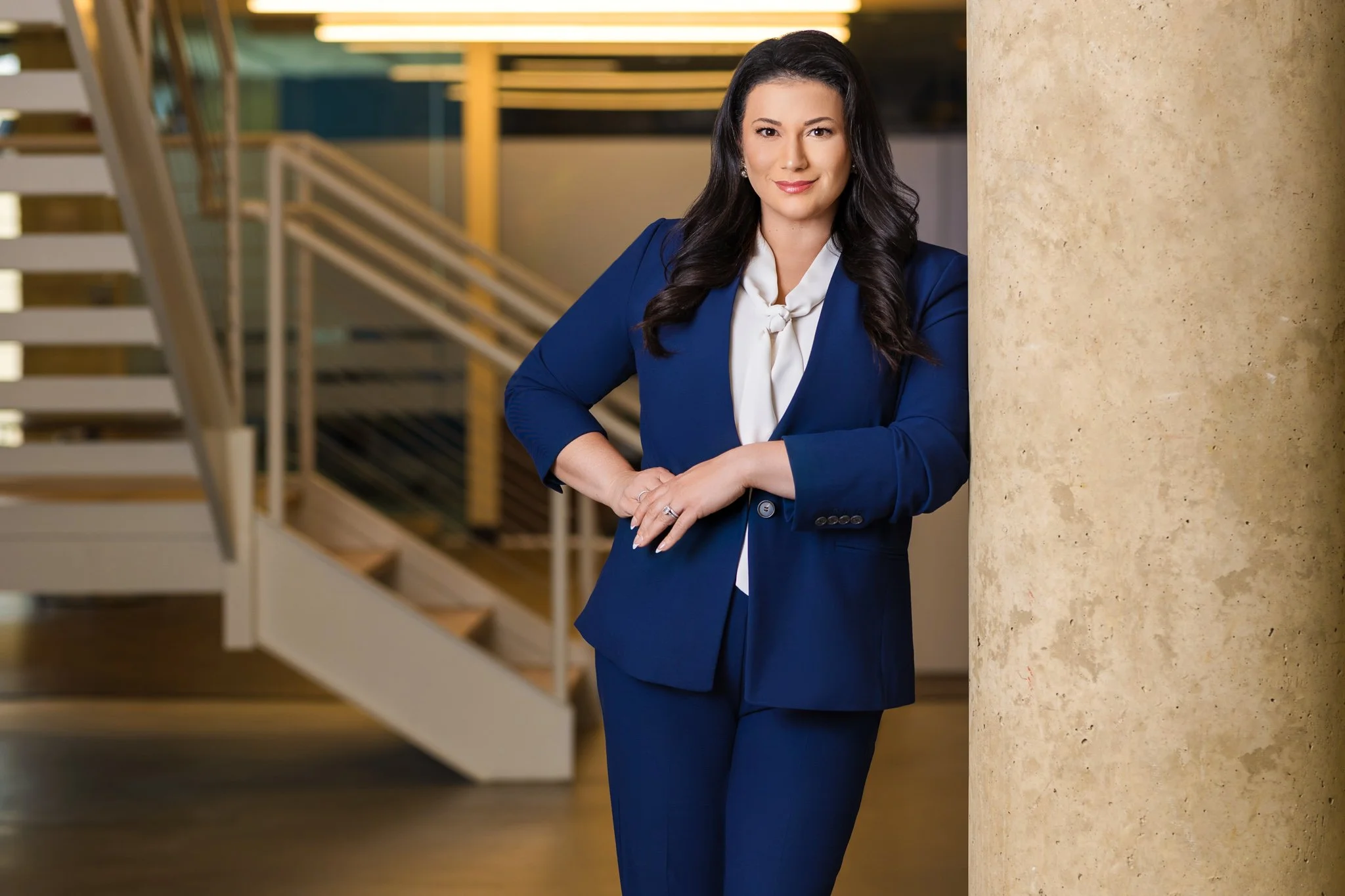 A woman in a blue business suit leaning against a beige column in a modern office building.