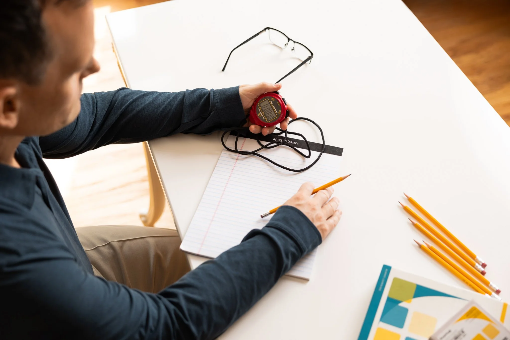 A person sitting at a white table with a stopwatch, reading glasses, a notepad, several yellow pencils, and pamphlets.
