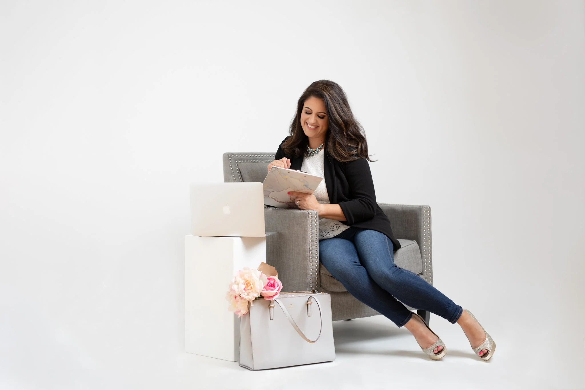 A woman sitting on a gray armchair, smiling, looking at a notebook, with a silver laptop on a white stand beside her, and a white handbag with flowers and a purse underneath, against a plain white background.
