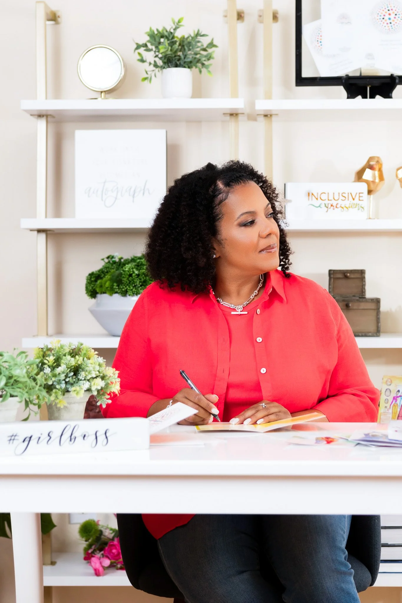A woman with curly hair sitting at a desk, wearing a red shirt and holding a pen, in an office or creative space with plants and decorative items in the background.