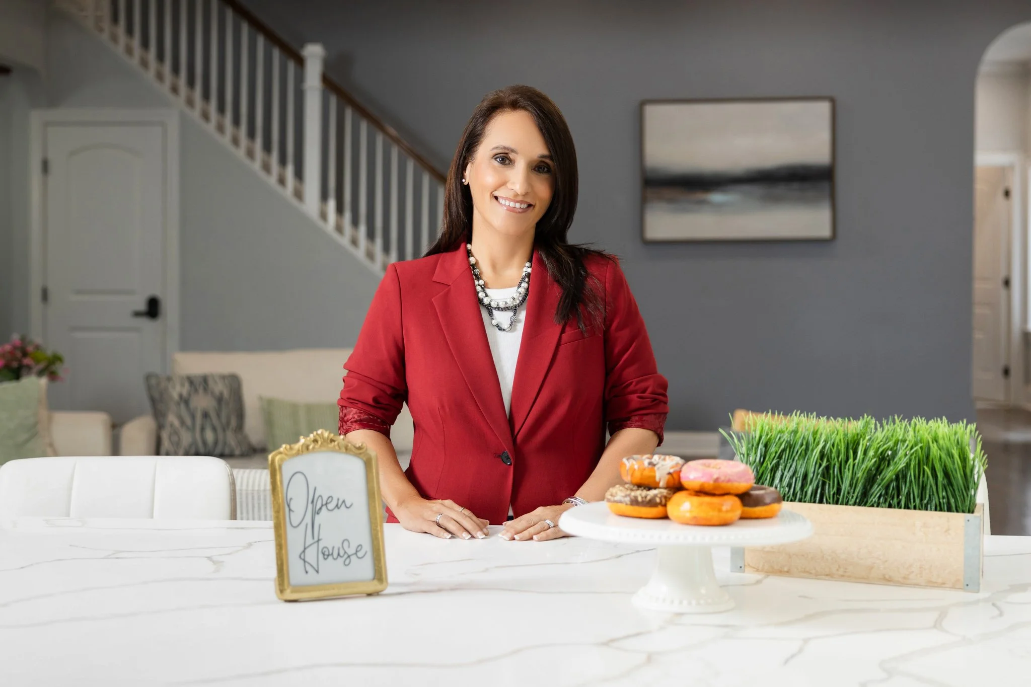 A woman in a red blazer stands behind a white kitchen island with a platter of colorful donuts, a small sign saying 'Open House', and a container of green grass plants in a bright, modern home.