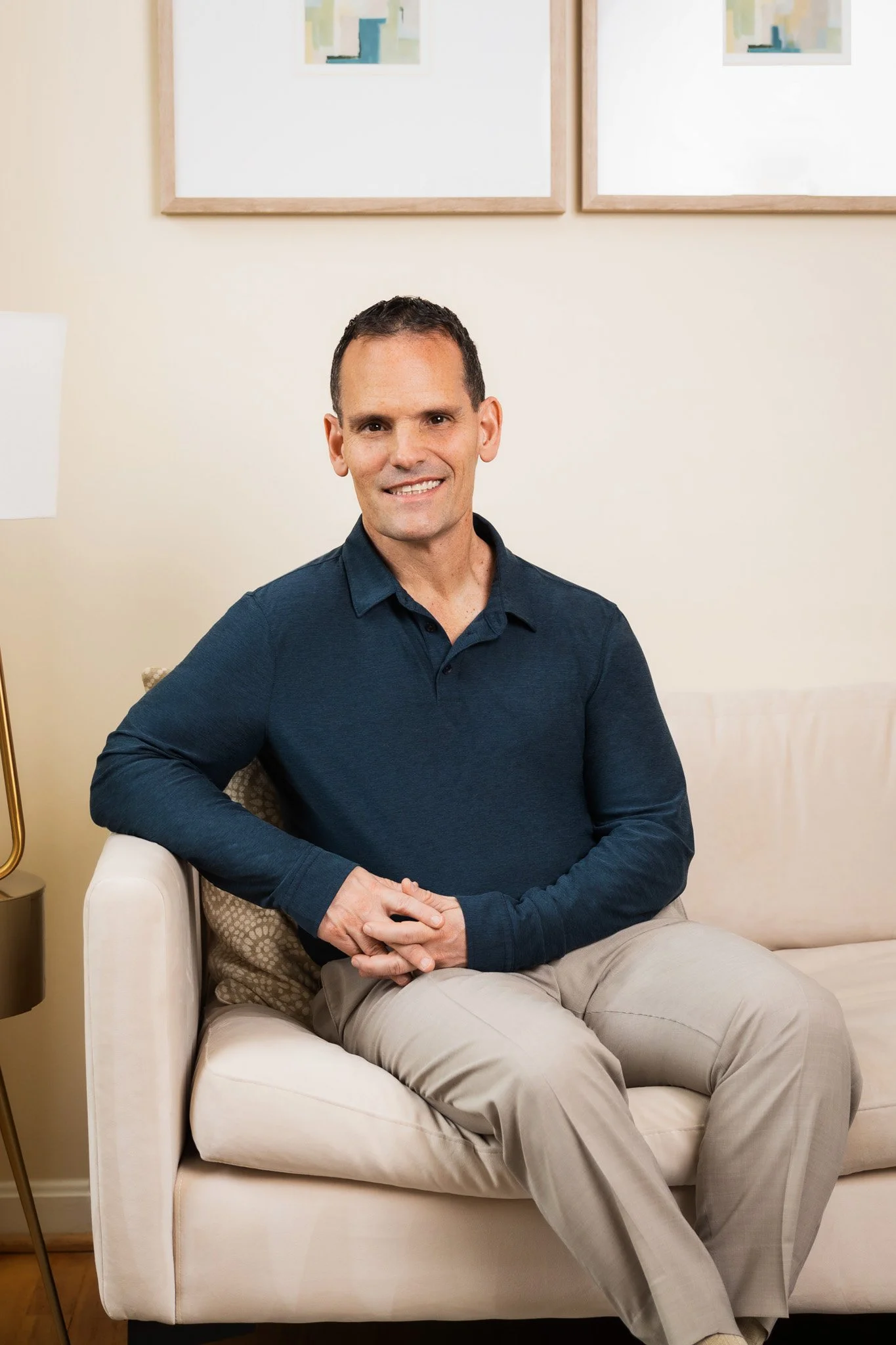 A man sitting on a beige sofa in a living room, wearing a dark blue long-sleeve shirt and beige pants, smiling at the camera. Behind him are framed abstract artwork and a cream-colored wall.