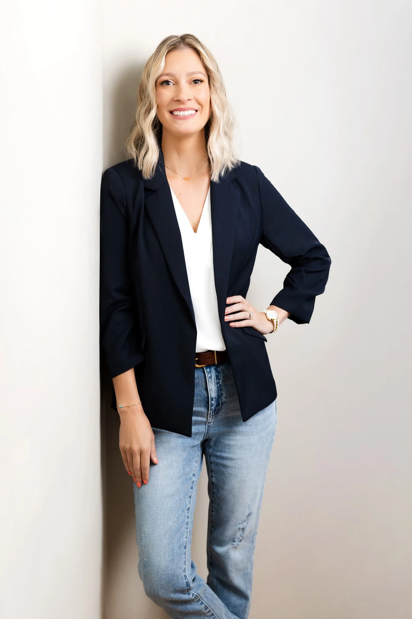 A woman with blonde hair, smiling, wearing a black blazer, white blouse, and light blue jeans, standing against a white wall.