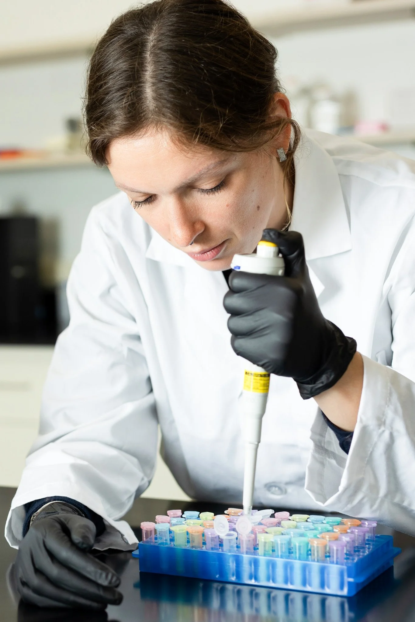 A female scientist wearing a white lab coat and black gloves, using a pipette to transfer liquid into test tubes arranged in a rack.