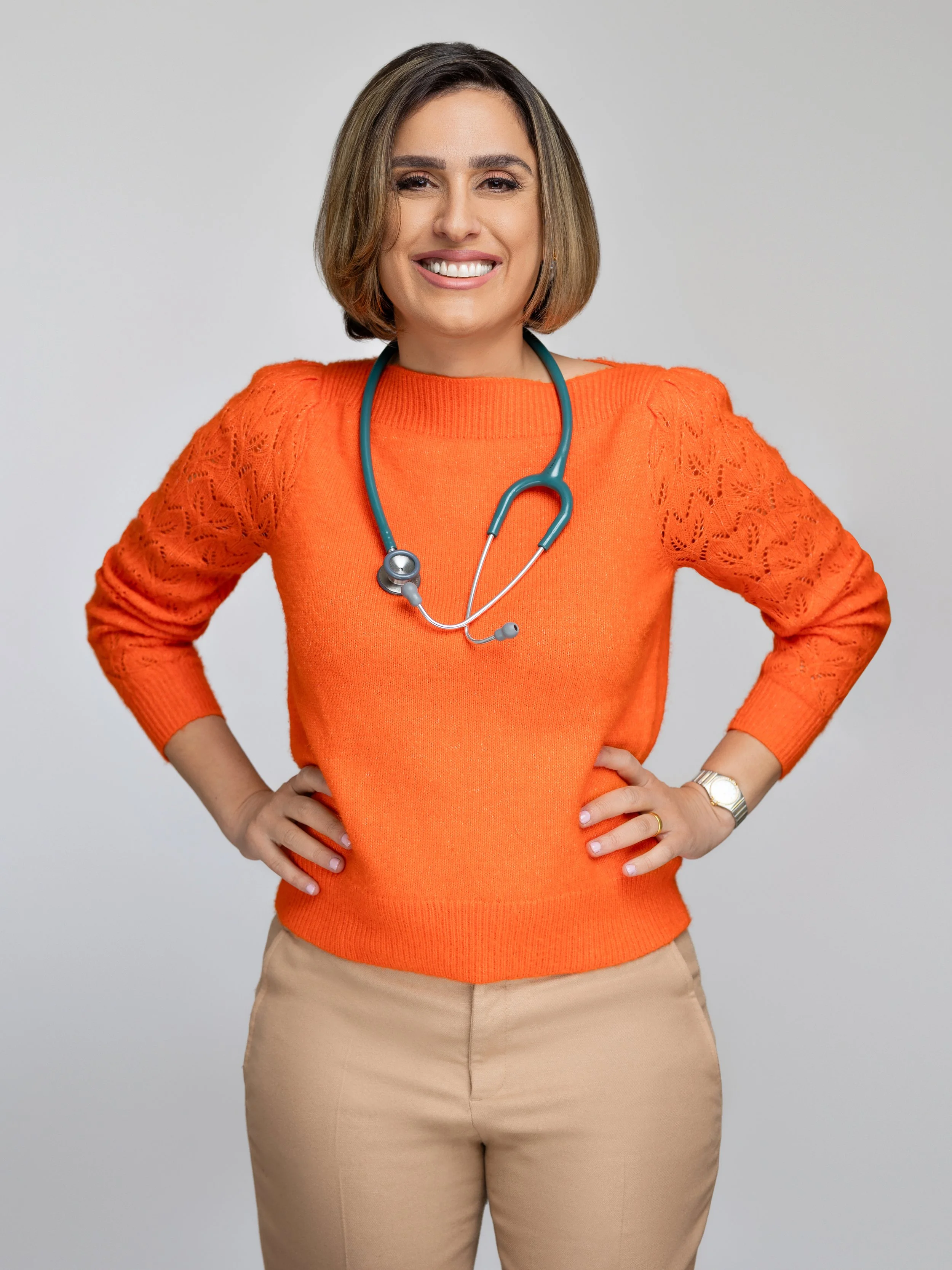 A smiling woman in an orange sweater with lace sleeves, beige pants, and a gold watch, standing with hands on hips and wearing a stethoscope around her neck.