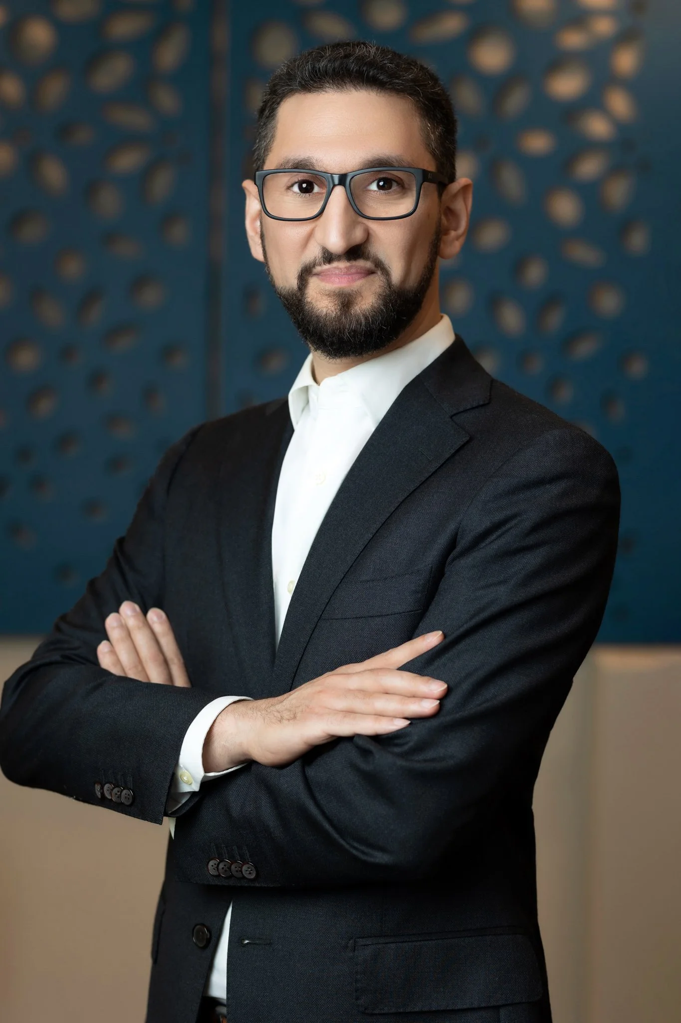 A man in a black suit with glasses and a beard standing with arms crossed in front of a blue decorative background.