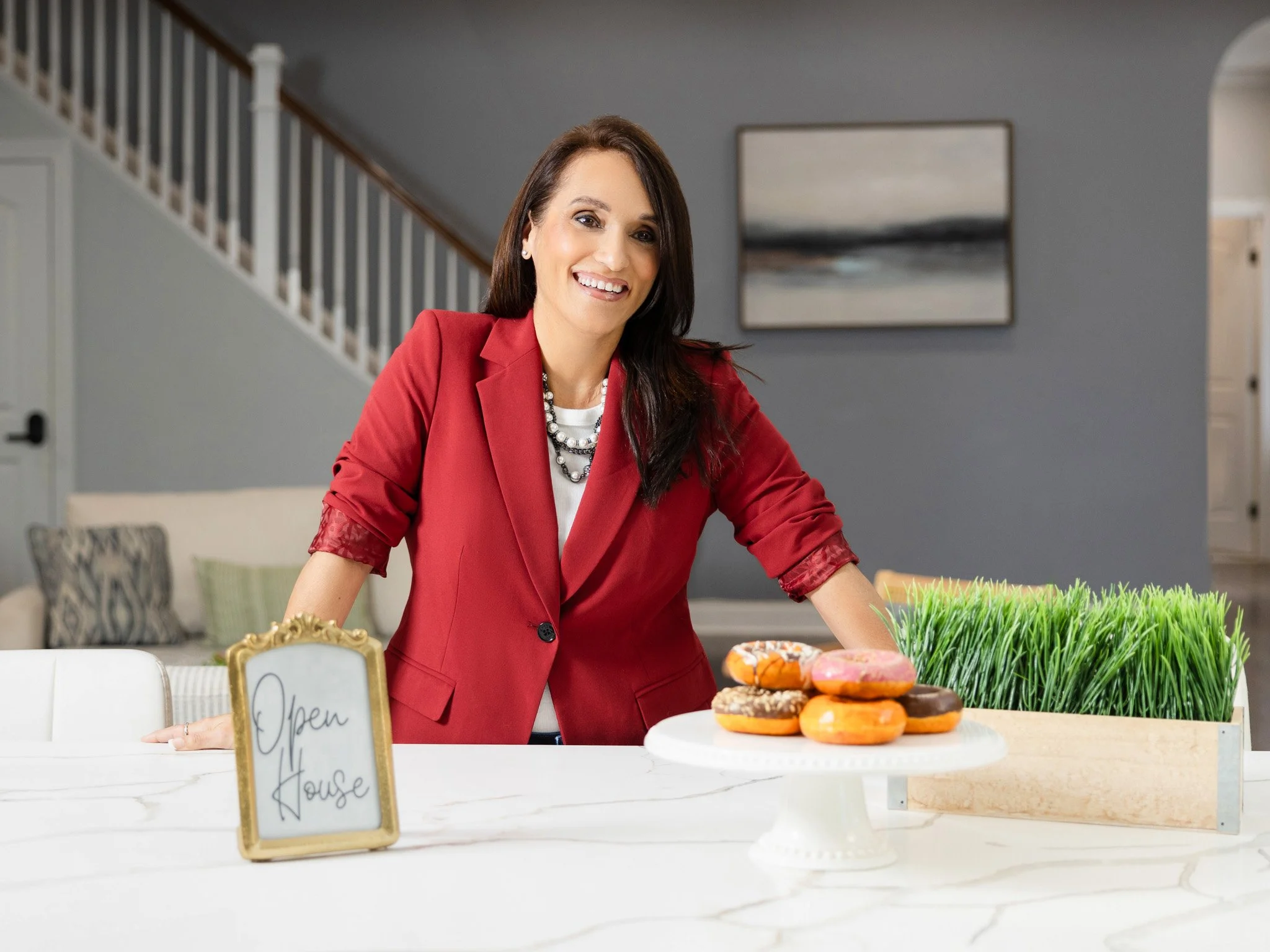 Woman in red blazer smiling at a table with donuts and a small sign that says 'Open House'