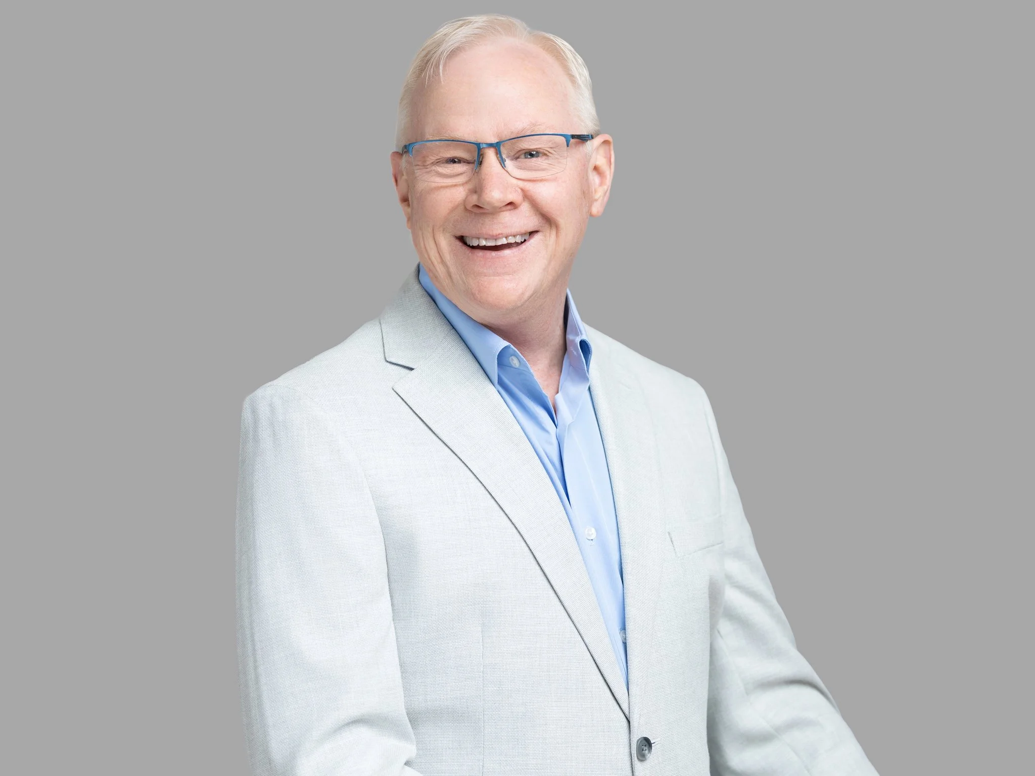 A smiling man with glasses, wearing a light grey suit and blue shirt, against a plain grey background.