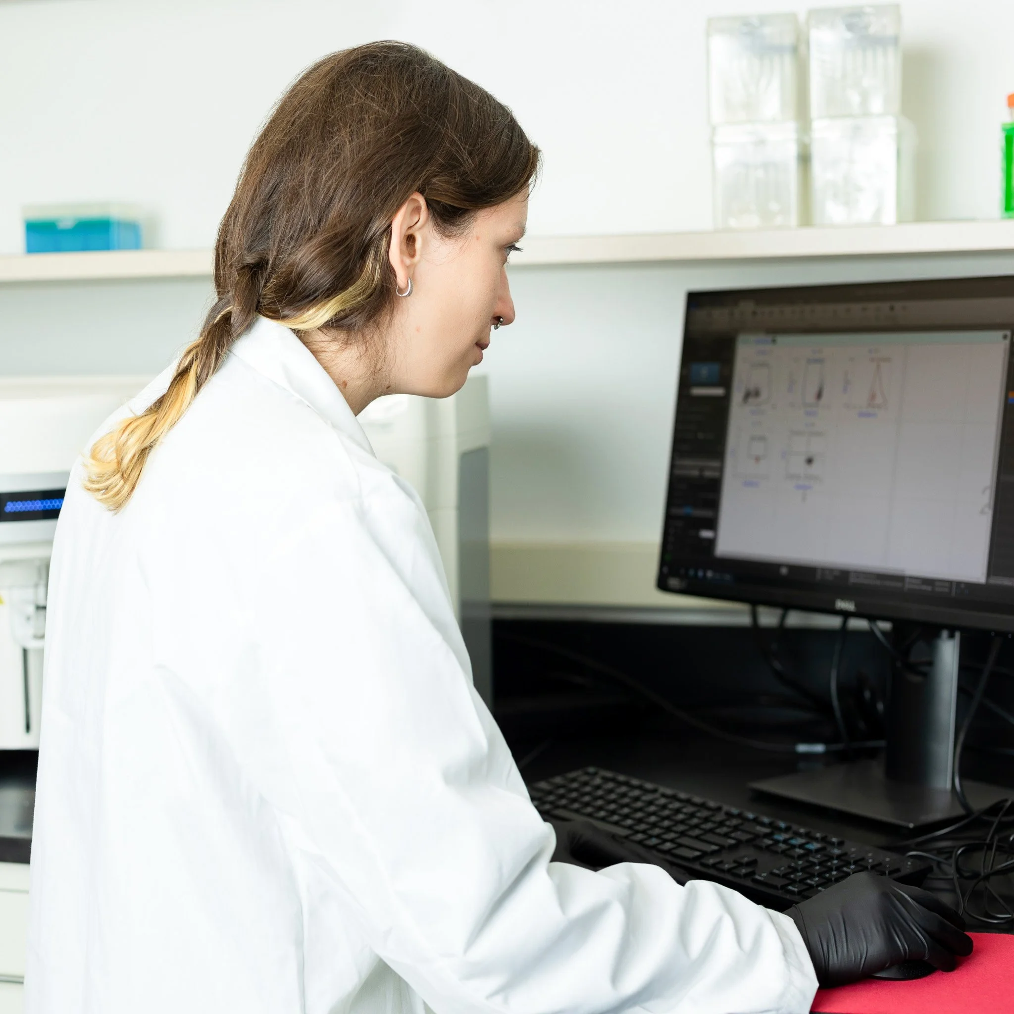 A female scientist wearing a white lab coat and black gloves working on a computer in a laboratory.