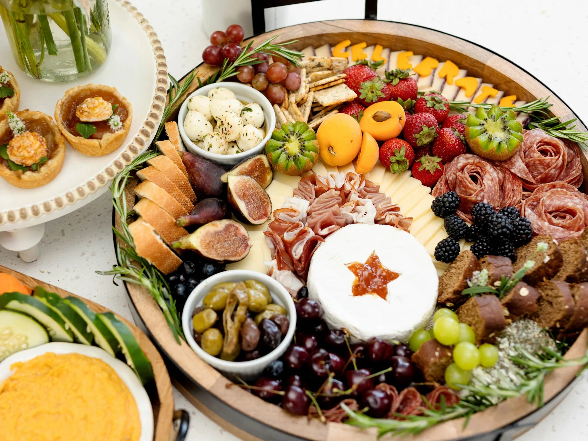 A charcuterie and cheese platter with an assortment of strawberries, blueberries, grapes, blackberries, cheeses, cured meats, figs, olives, and crackers, garnished with herbs.