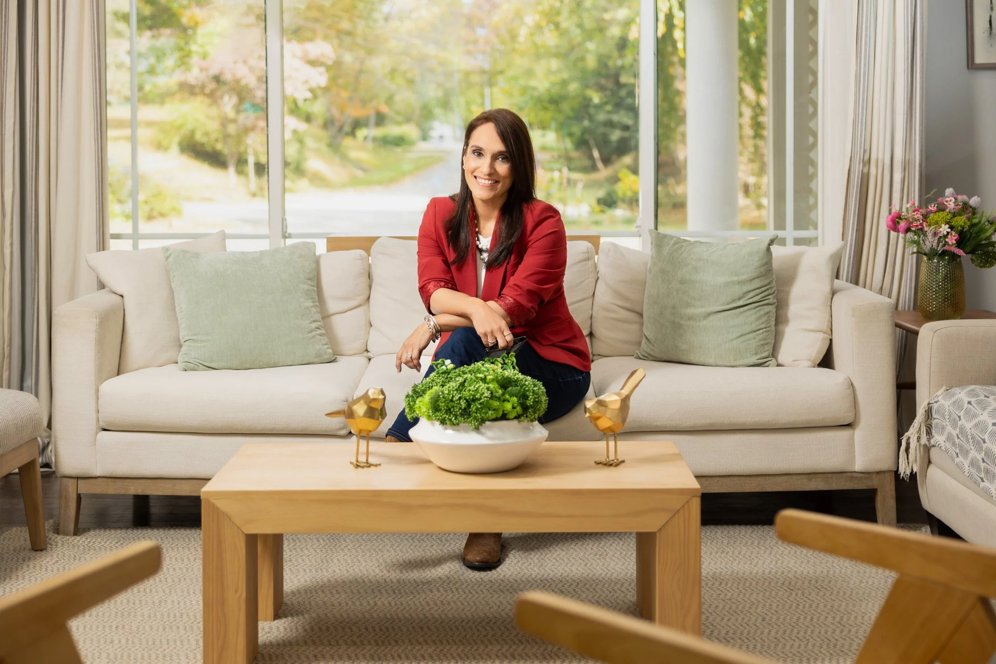 A woman in a red blazer sitting on a beige sofa with green pillows in a bright living room with a large window behind her showing greenery outside. A wooden coffee table in front of her has a white bowl of green plants and gold bird figurines. There 
