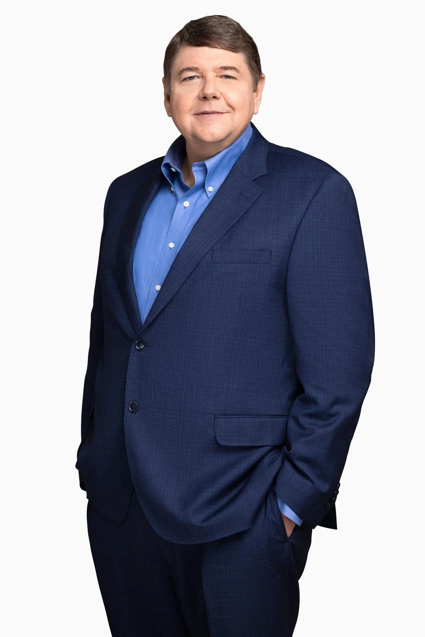 A man in a navy suit with a blue shirt standing against a white background.
