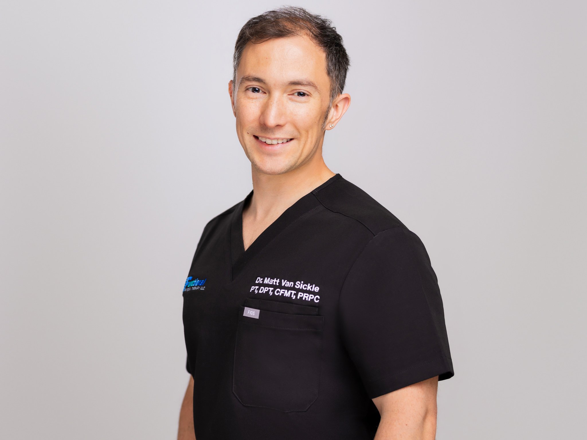 A smiling man dressed in black scrubs smiling at the camera. The background is light gray.