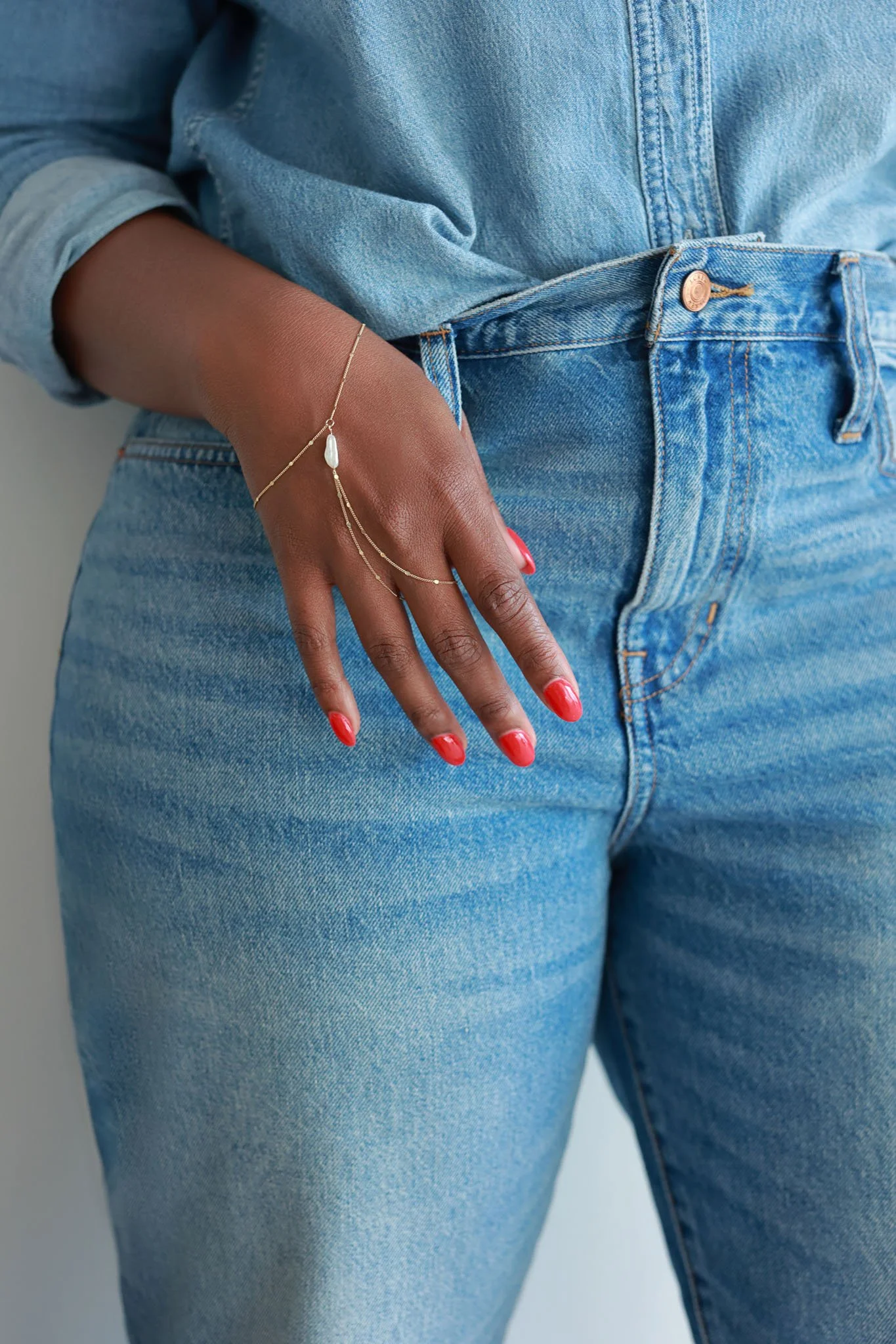Close-up of a person wearing a denim shirt and jeans, with their hand resting on their hip, accessorized with delicate gold jewelry and red nail polish.