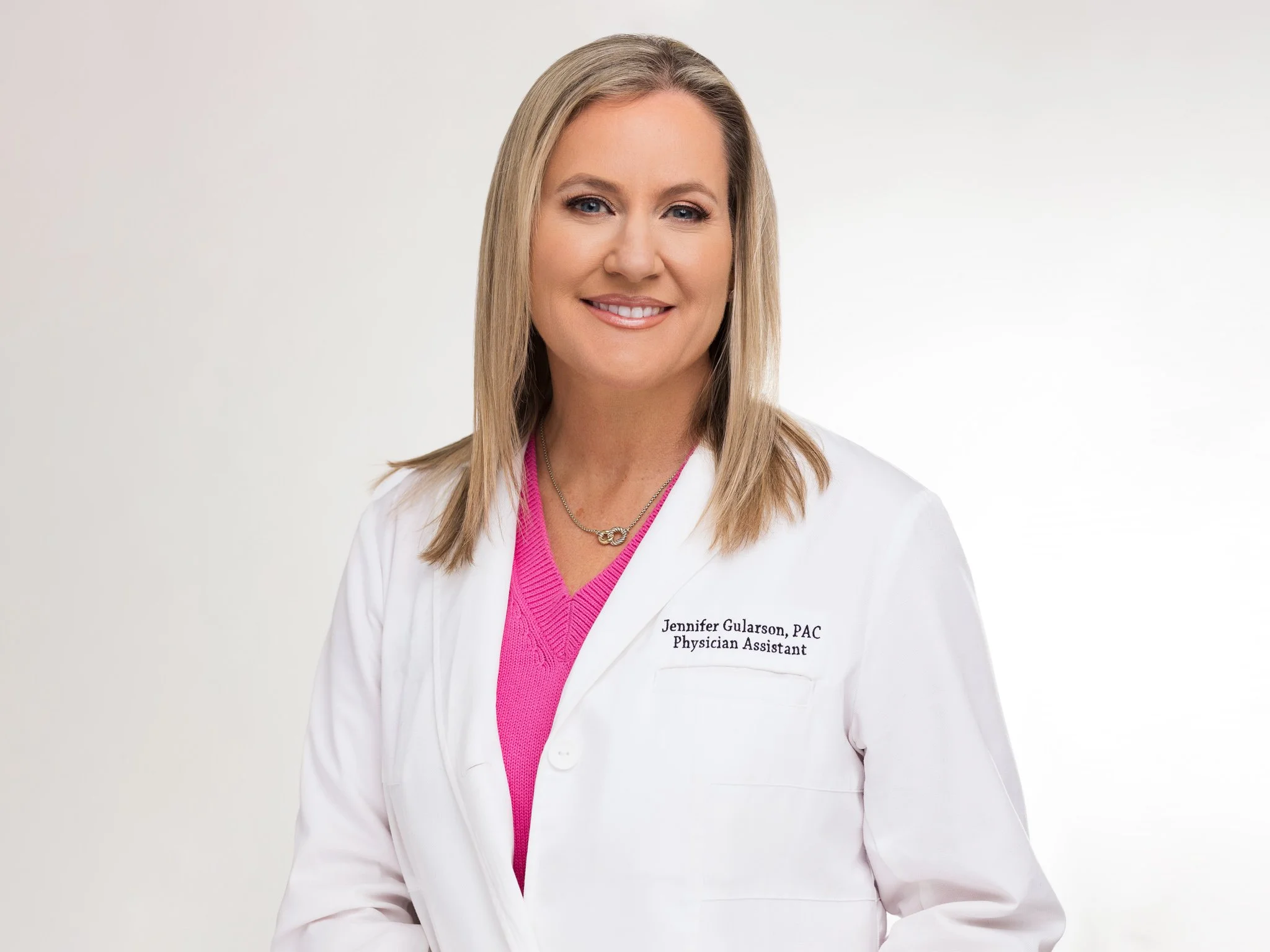 A woman with shoulder-length blonde hair wearing a white lab coat over a pink sweater, smiling at the camera, standing against a plain white background.