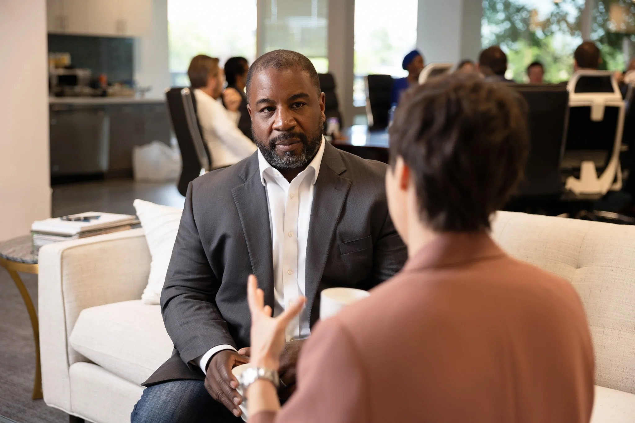 A man in a gray suit is speaking with a woman in a brown blazer in a professional office setting, with other people working at desks in the background.