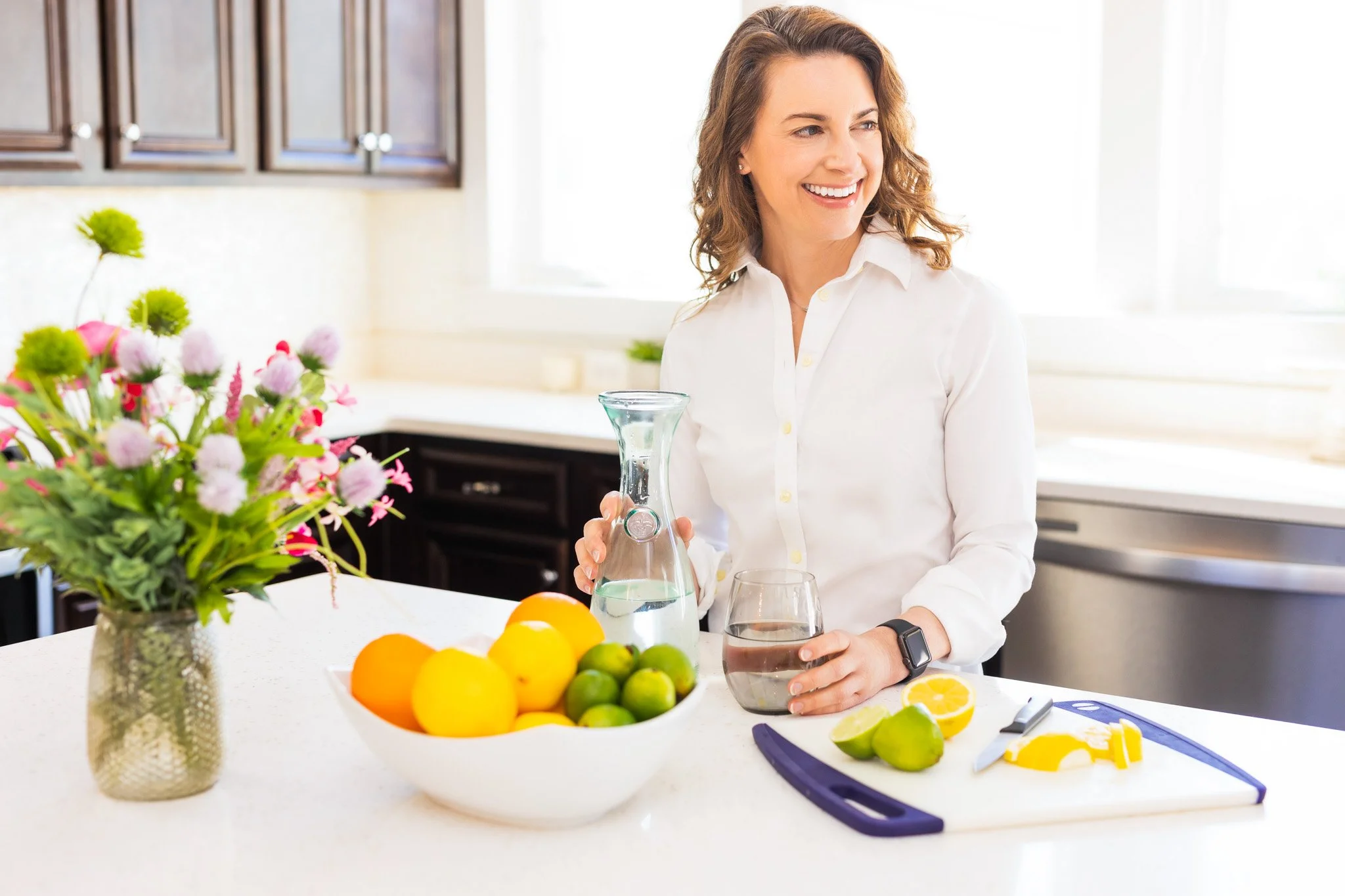 happy smiling personal wellness coach leaning on a kitchen island with a glass carafe of water and vibrant fruit