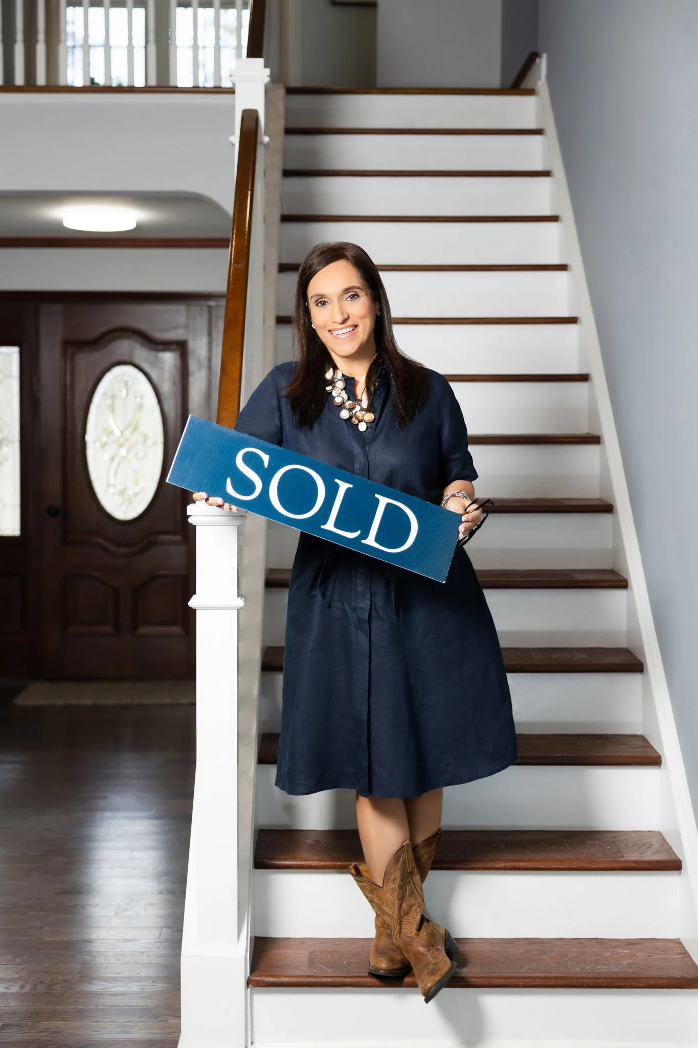 Woman standing on staircase holding a sign that reads 'SOLD,' inside a house.