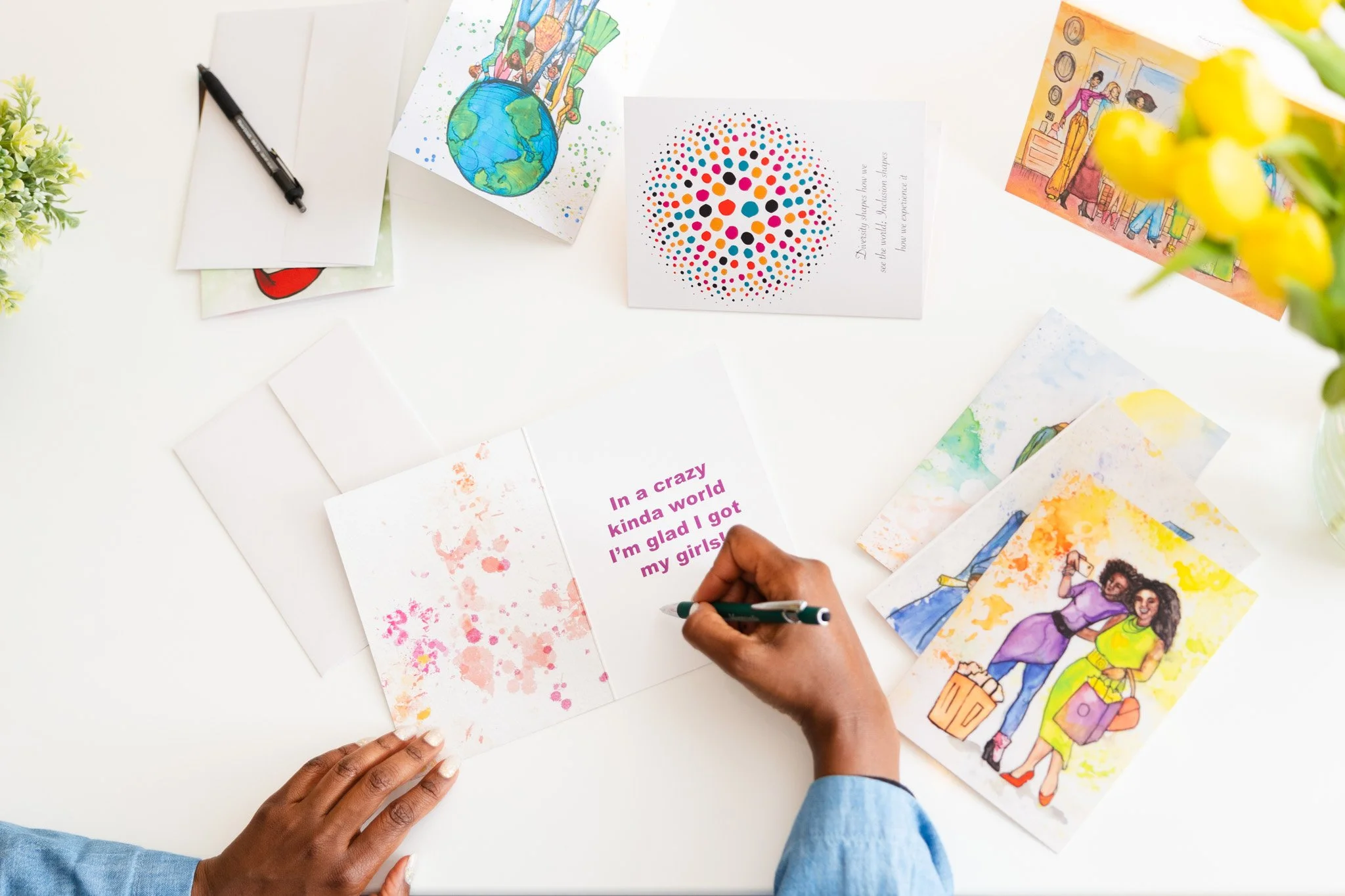 A person writes on a card with pink, purple, and orange text that reads, 'In a crazy kinda world I'm glad I got my girls!' surrounded by colorful greeting cards and watercolor illustrations on a white table.